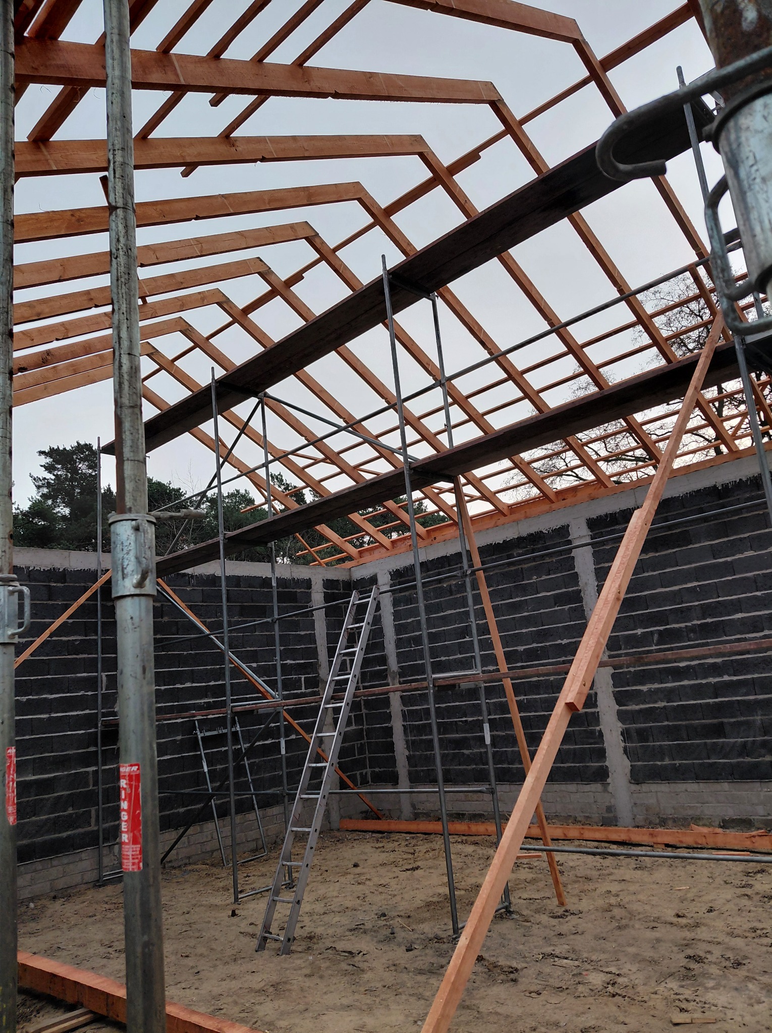 Wznoszenie drewnianej konstrukcji dachu z widocznymi elementami więźby, rusztowaniami i ścianami z bloczków betonowych. Widok z dołu na budowę w toku.