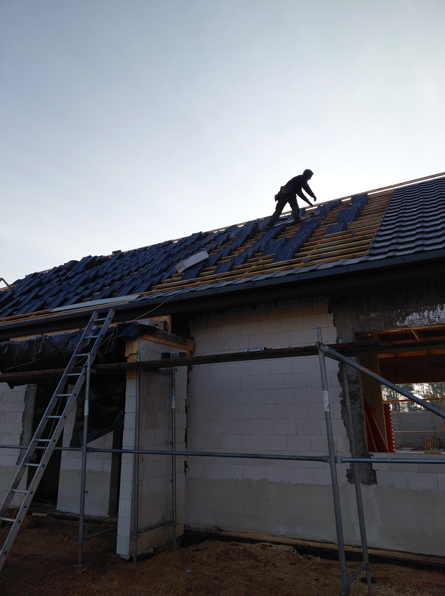 Dekarz w trakcie krycia dachu na nowym budynku. Widoczne rusztowania, drabina i częściowo pokryty dach z czarną folią i dachówką. Ujęcie z dołu.
