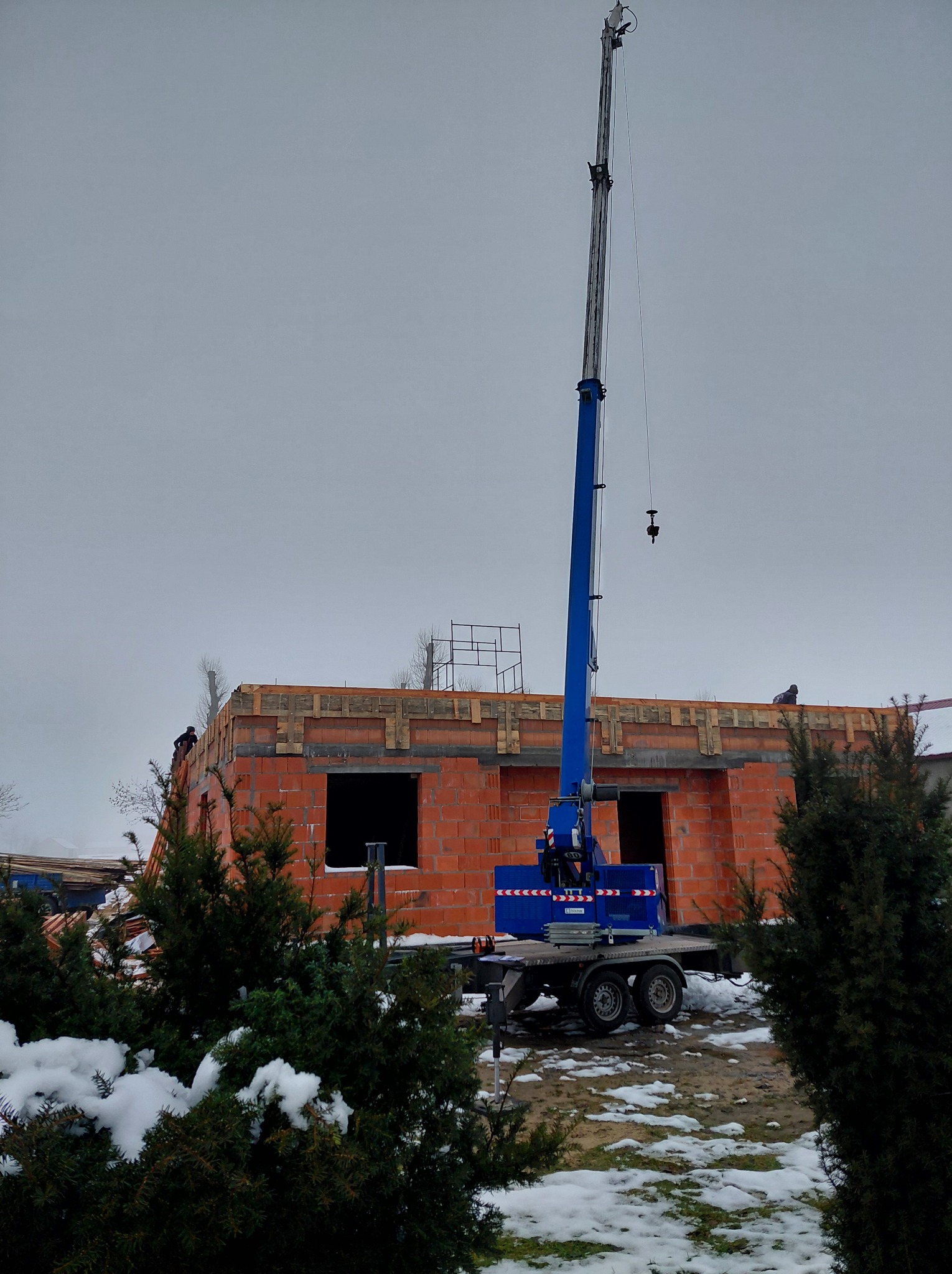 Budowa domu z czerwonej cegły w zimowej scenerii. Na pierwszym planie zielone krzewy ze śniegiem, w tle niebieski dźwig i pracujący dekarze na dachu.