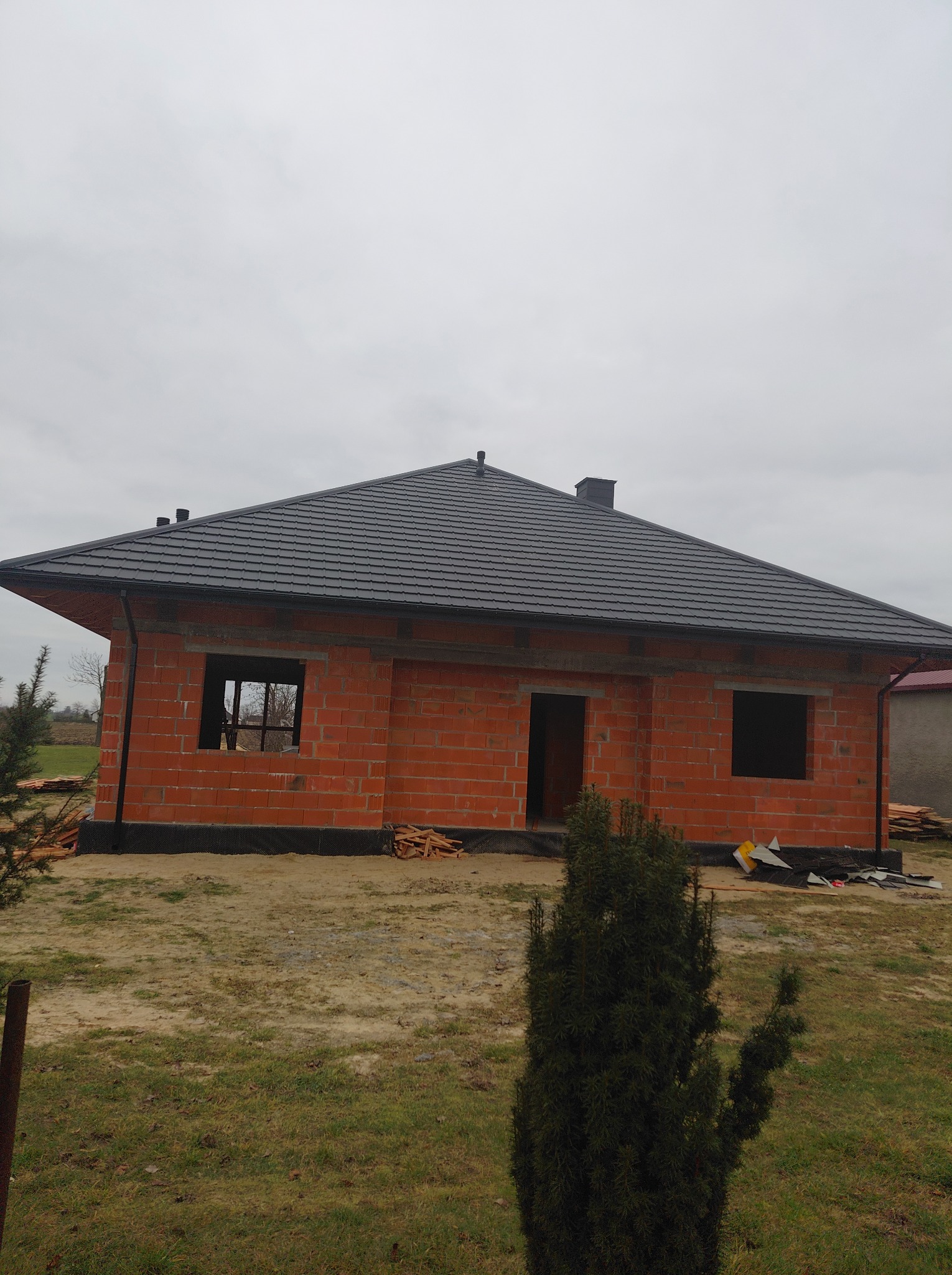 Dom jednorodzinny w trakcie budowy, z ciemnoszarą dachówką i widocznymi elementami konstrukcyjnymi, na tle pochmurnego nieba. Widoczne rynny i obróbki blacharskie.