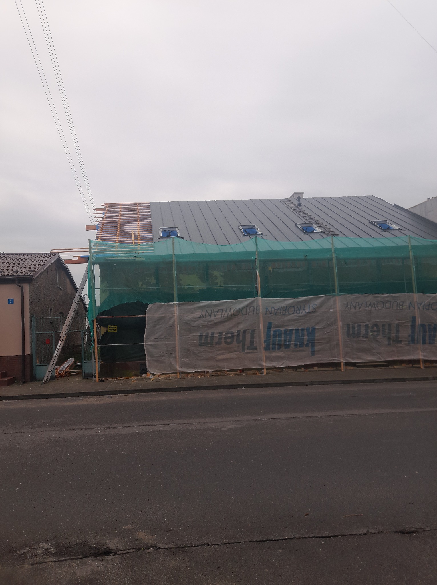 Dom w trakcie wymiany dachu z blachy, zabezpieczony siatką i folią budowlaną. Widoczne elementy więźby dachowej i okna dachowe. Prace dekarskie w toku.