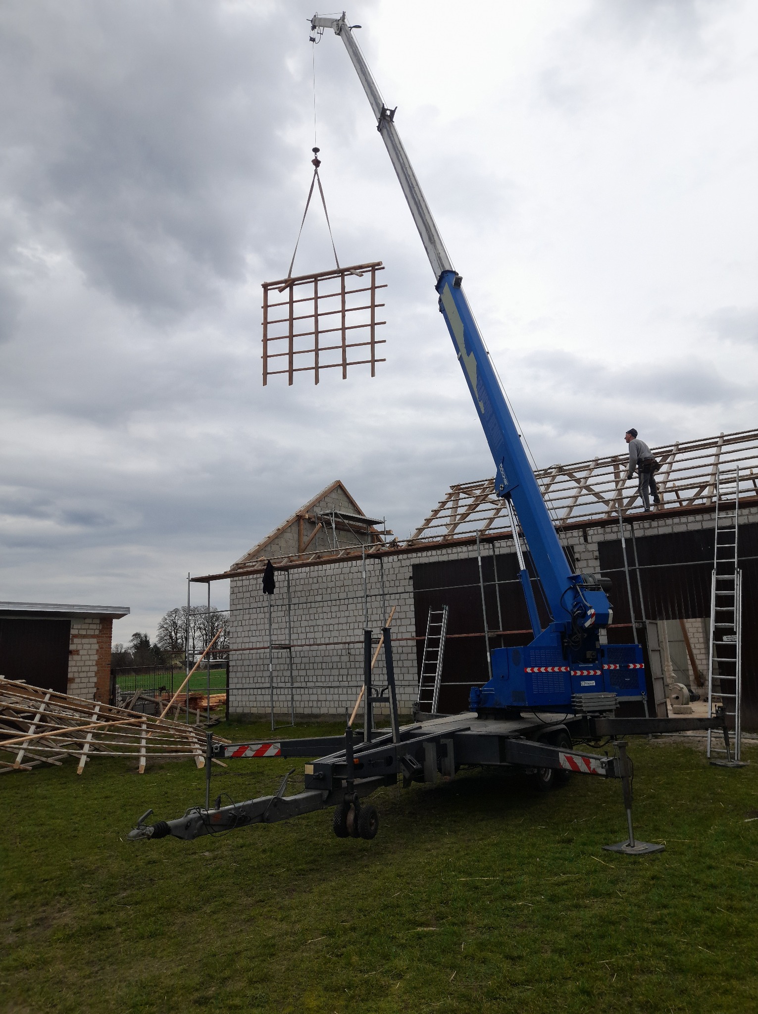 Montaż więźby dachowej za pomocą dźwigu na budowie. Widoczny pracownik na dachu i elementy konstrukcyjne. Pochmurne niebo w tle.