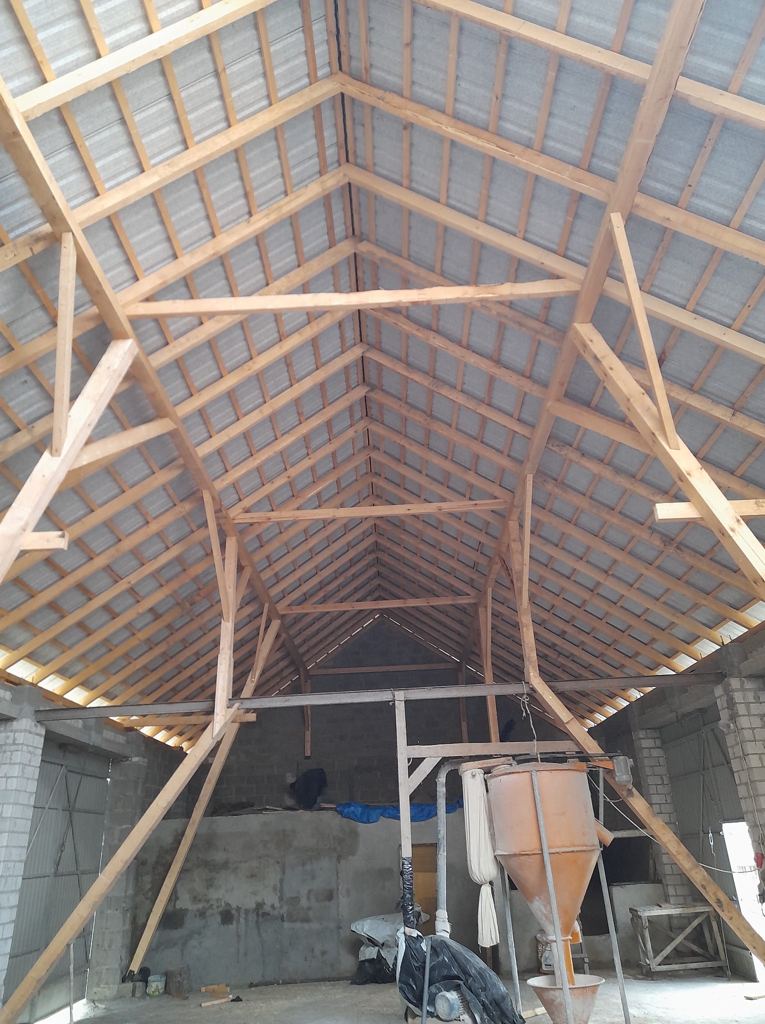 Wnętrze budynku z widoczną drewnianą konstrukcją więźby dachowej i szarym pokryciem. Widok z dołu na elementy dachu w trakcie budowy.
