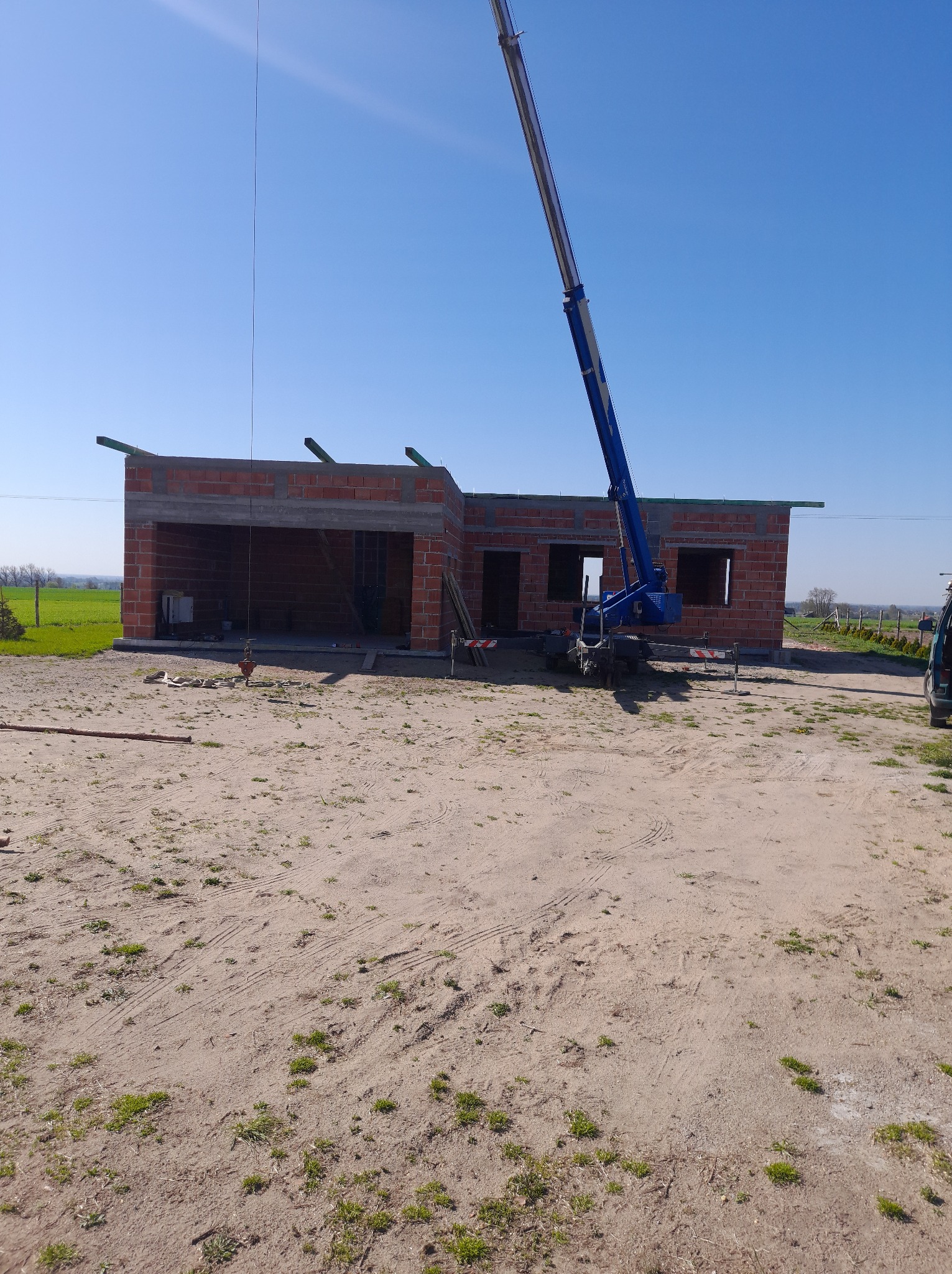 Budowa domu jednorodzinnego w stanie surowym otwartym, z widocznym dźwigiem budowlanym na tle błękitnego nieba. Ceglana konstrukcja z elementami drewnianymi.