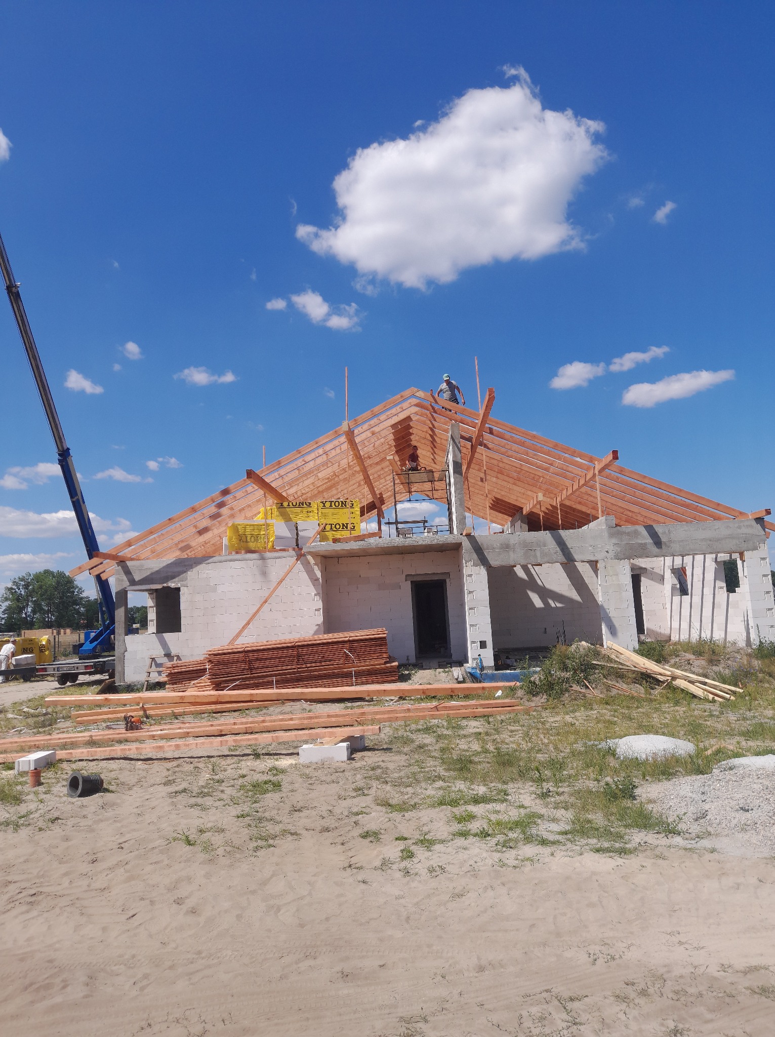 Budowa domu jednorodzinnego w trakcie wznoszenia więźby dachowej. Widoczny dźwig i pracownicy na dachu. W tle błękitne niebo z chmurami.