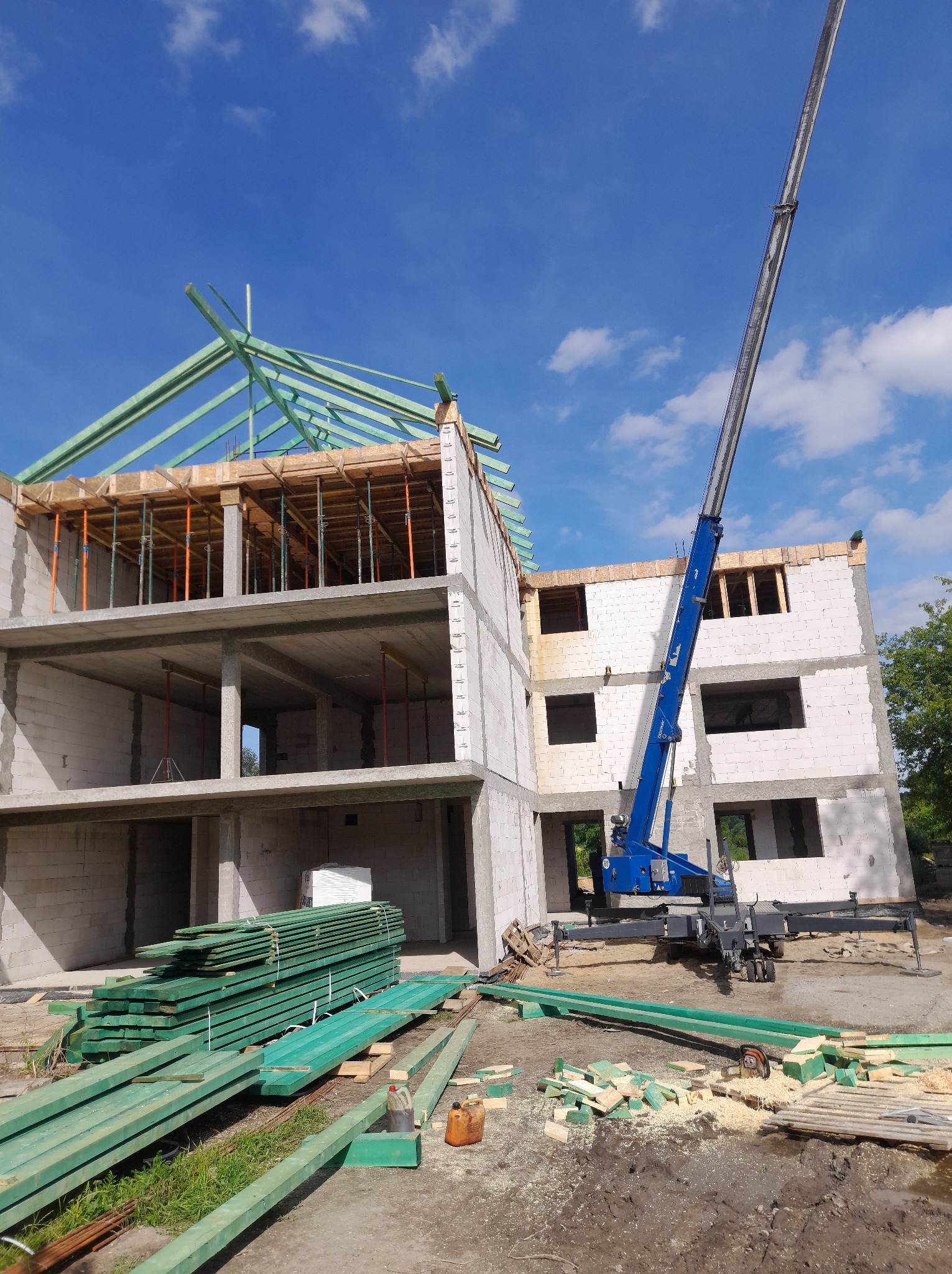 Budowa domu w stanie surowym otwartym z widoczną konstrukcją dachu i dźwigiem. Na pierwszym planie stos zielonych desek i resztki drewna po obróbce.