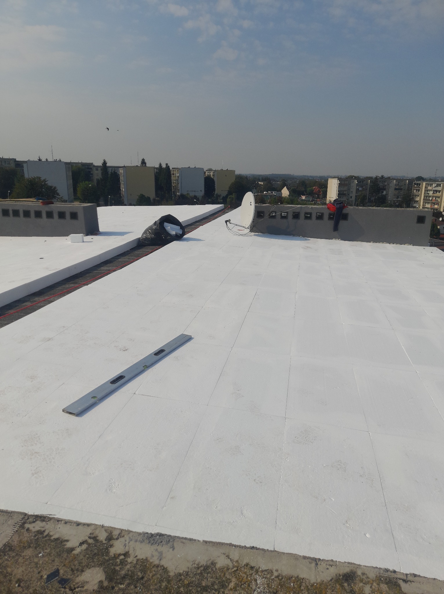 Białe płyty termoizolacyjne na płaskim dachu budynku, poziomica, worek z odpadami, antena satelitarna. W tle zabudowa miejska i błękitne niebo.