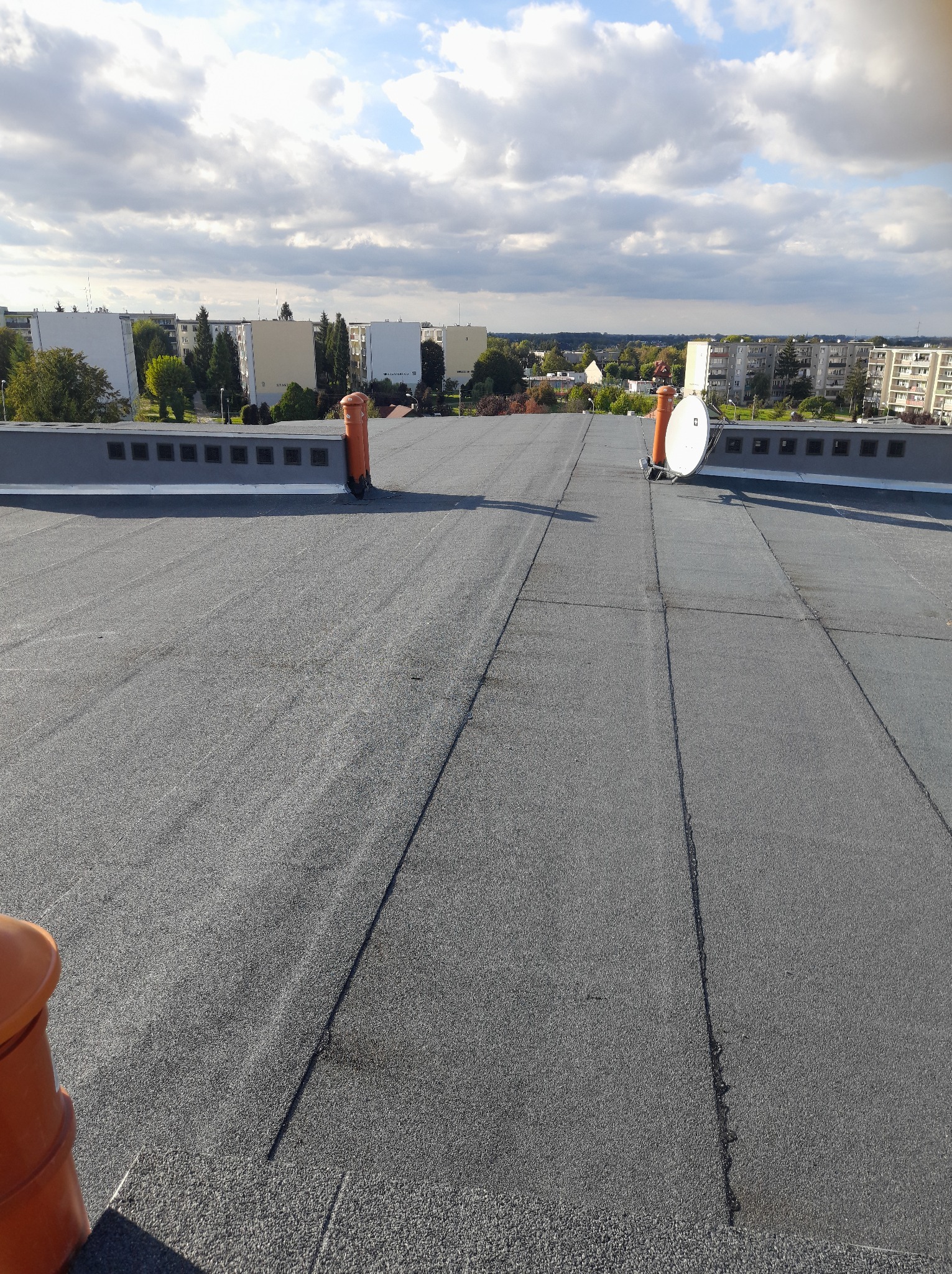 Szary, płaski dach pokryty papą termozgrzewalną z dwoma kominami wentylacyjnymi i anteną satelitarną na tle zabudowy miejskiej i pochmurnego nieba.