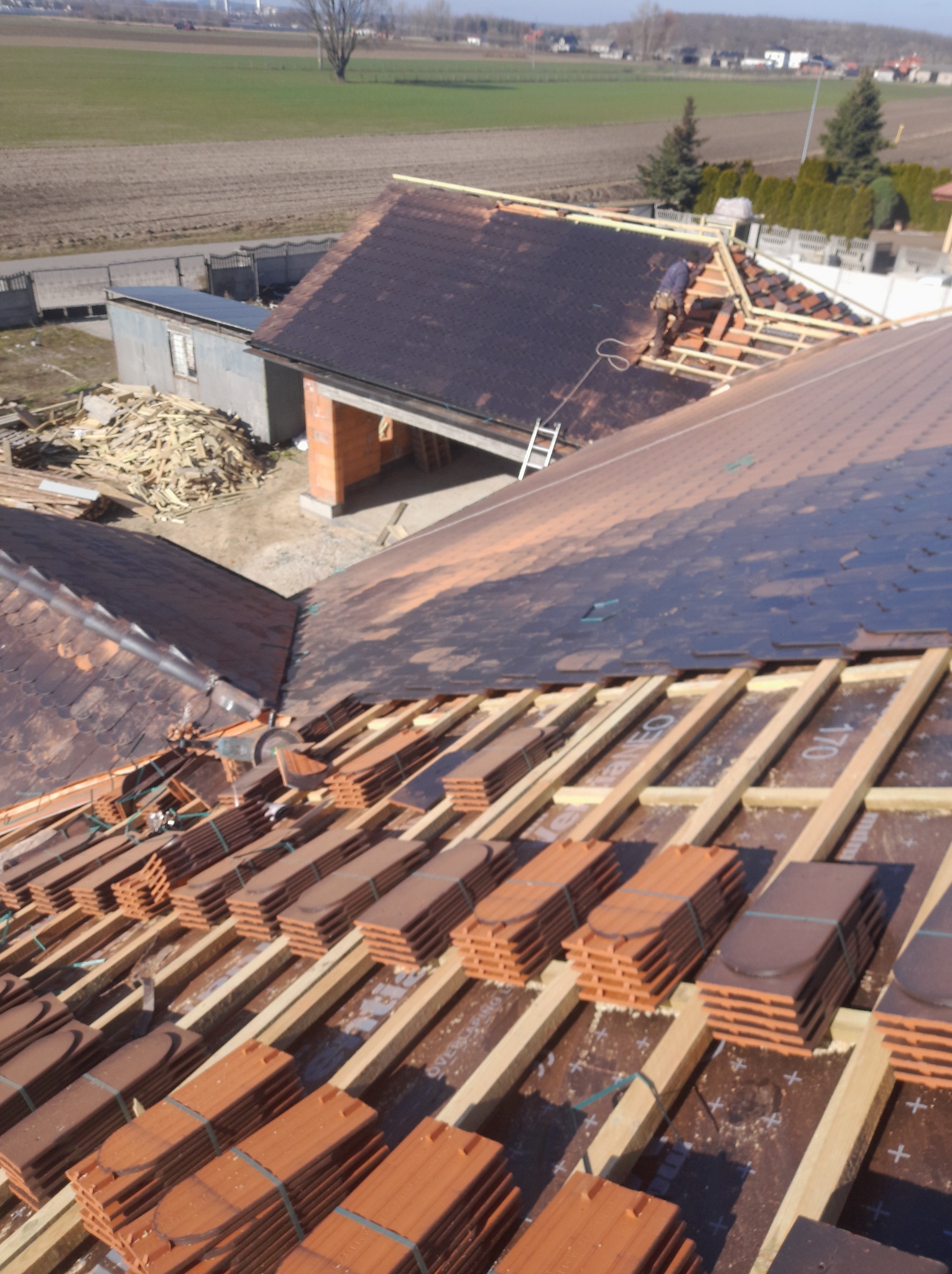 Układanie brązowej dachówki ceramicznej na drewnianej konstrukcji dachu. Widoczny dekarz pracujący na dachu domu jednorodzinnego w słoneczny dzień.