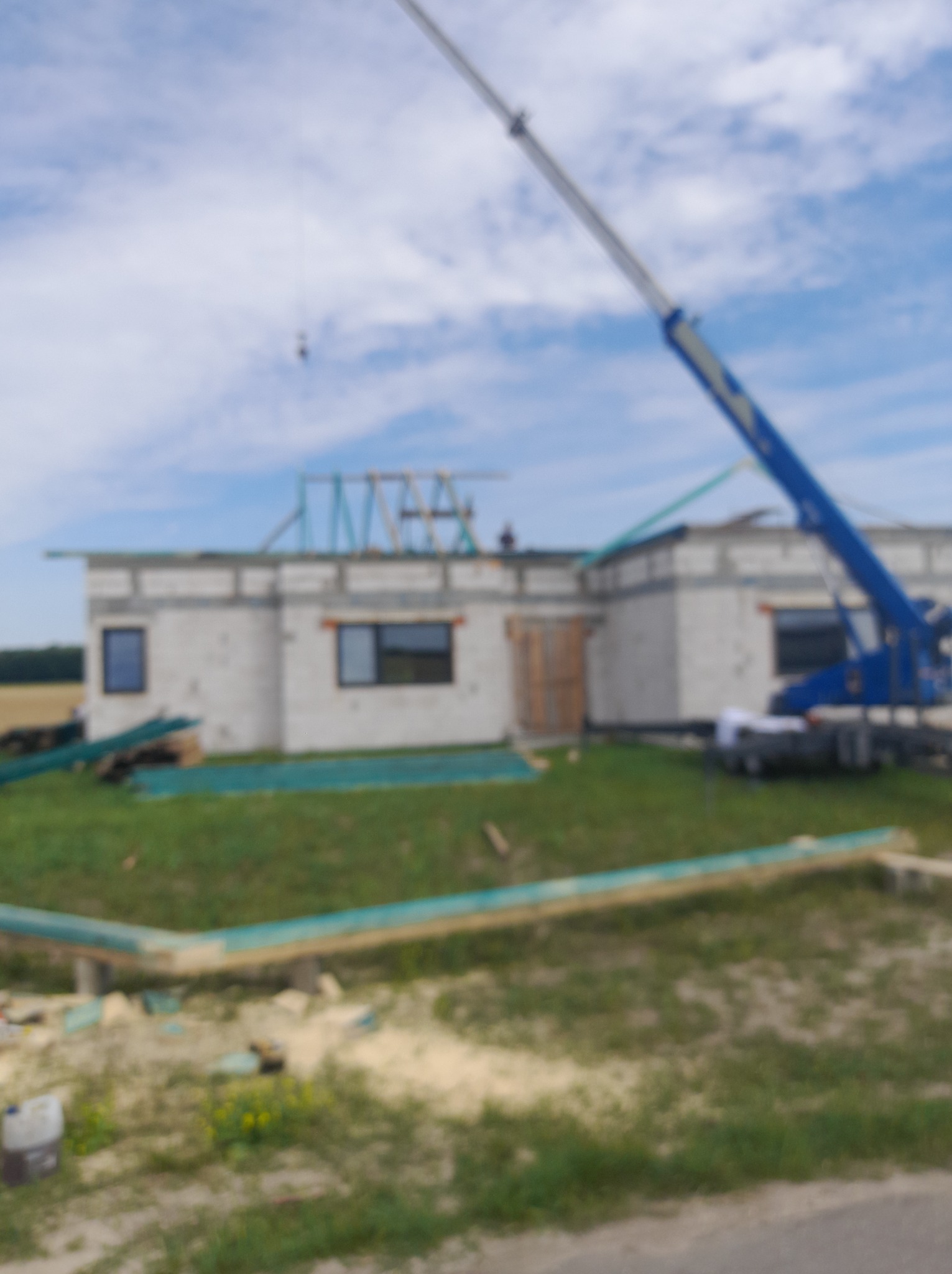 Budowa domu jednorodzinnego z widoczną konstrukcją więźby dachowej i dźwigiem. Elementy konstrukcyjne leżą na ziemi. Rozmyte tło.