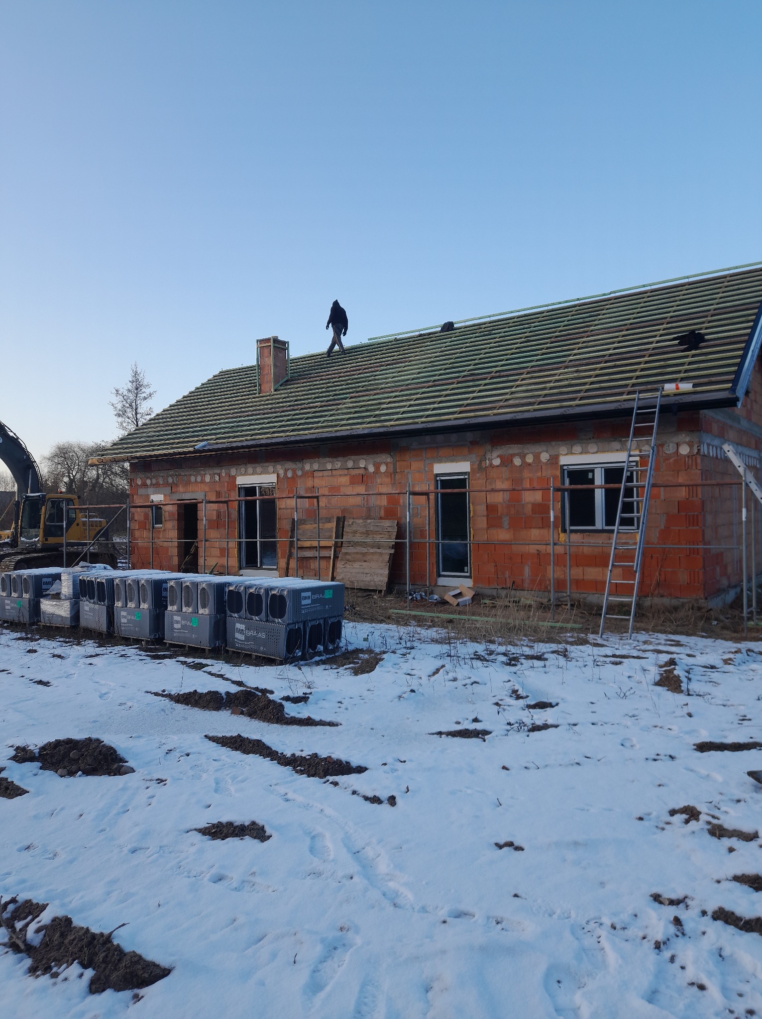 Budowa domu z częściowo pokrytym dachem dachówką ceramiczną. Na dachu stoi dekarz. Na pierwszym planie palety z dachówką. Zima.