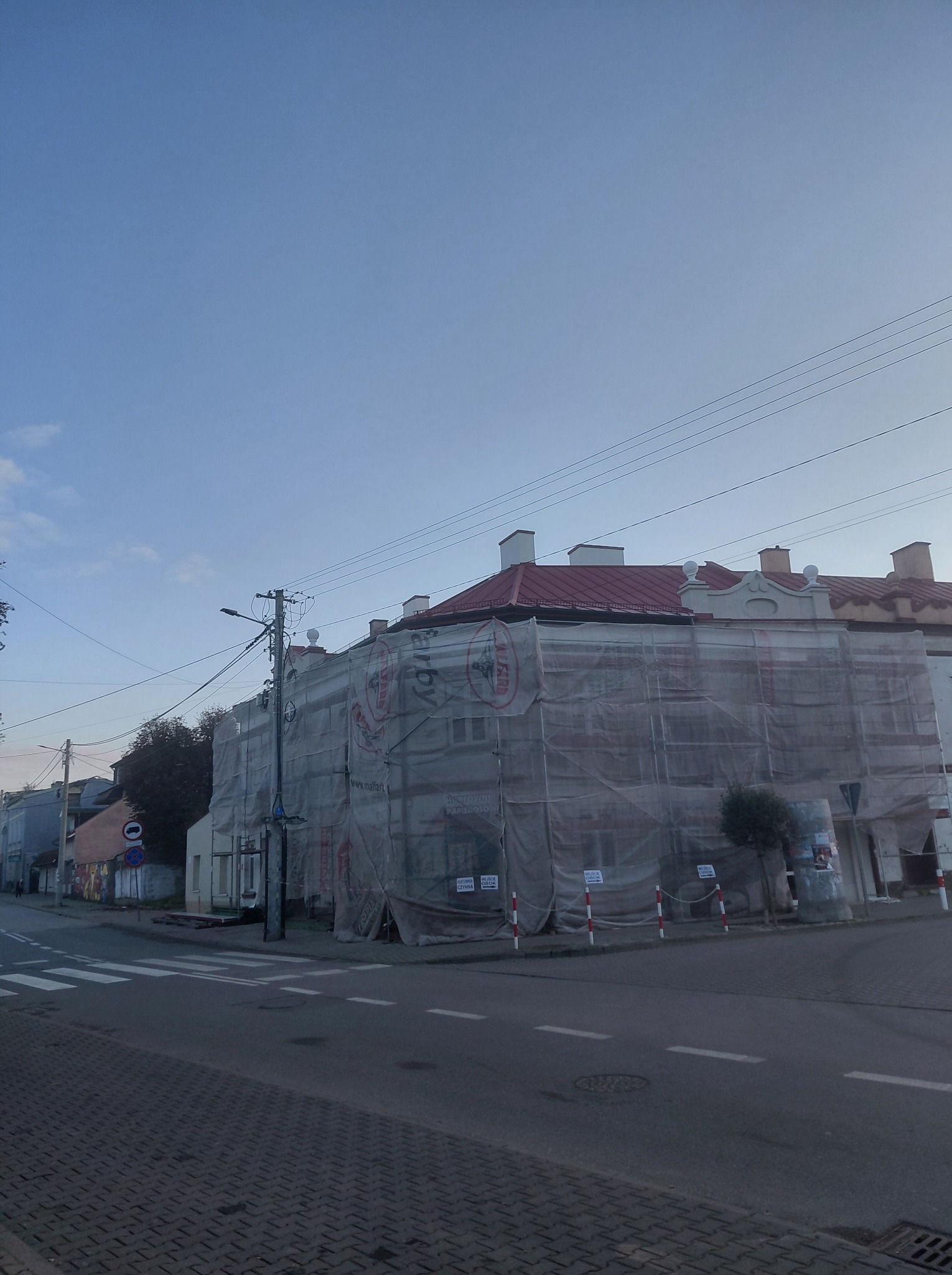 Budynek w trakcie renowacji, osłonięty siatką ochronną. Widoczny fragment dachu z czerwonej dachówki i elementy dekoracyjne fasady. Ujęcie z poziomu ulicy.