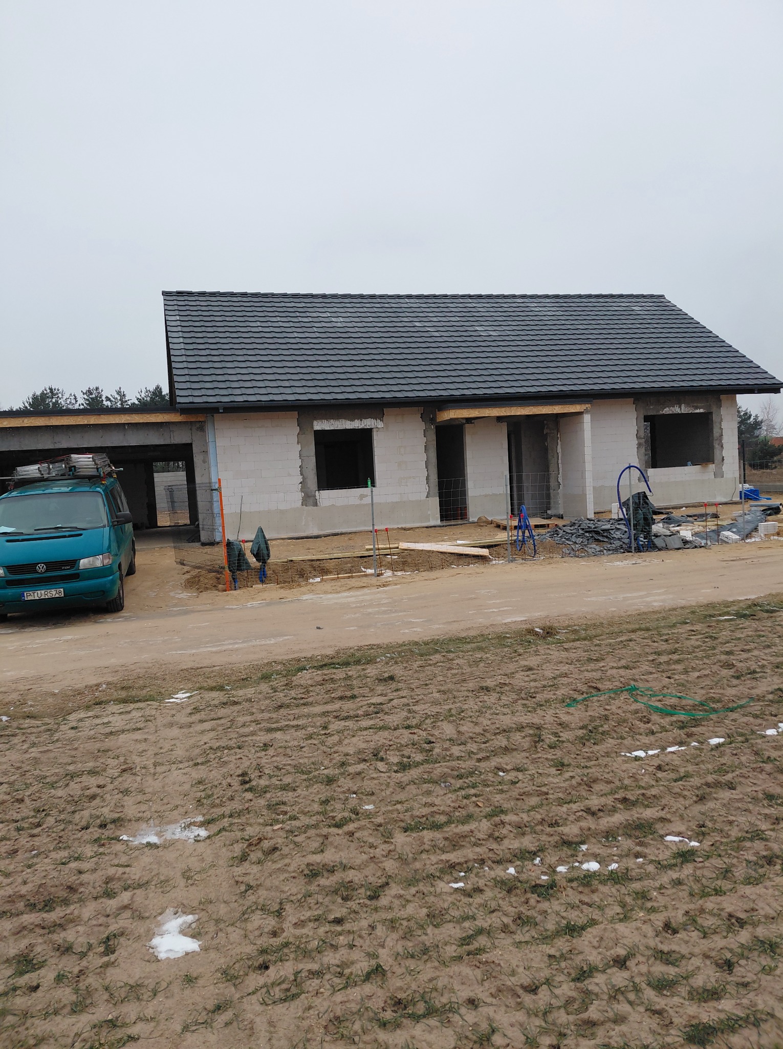 Nowy, grafitowy dach z dachówki ceramicznej na surowym budynku w trakcie budowy. Na pierwszym planie zielony bus z drabiną. Pochmurny dzień.