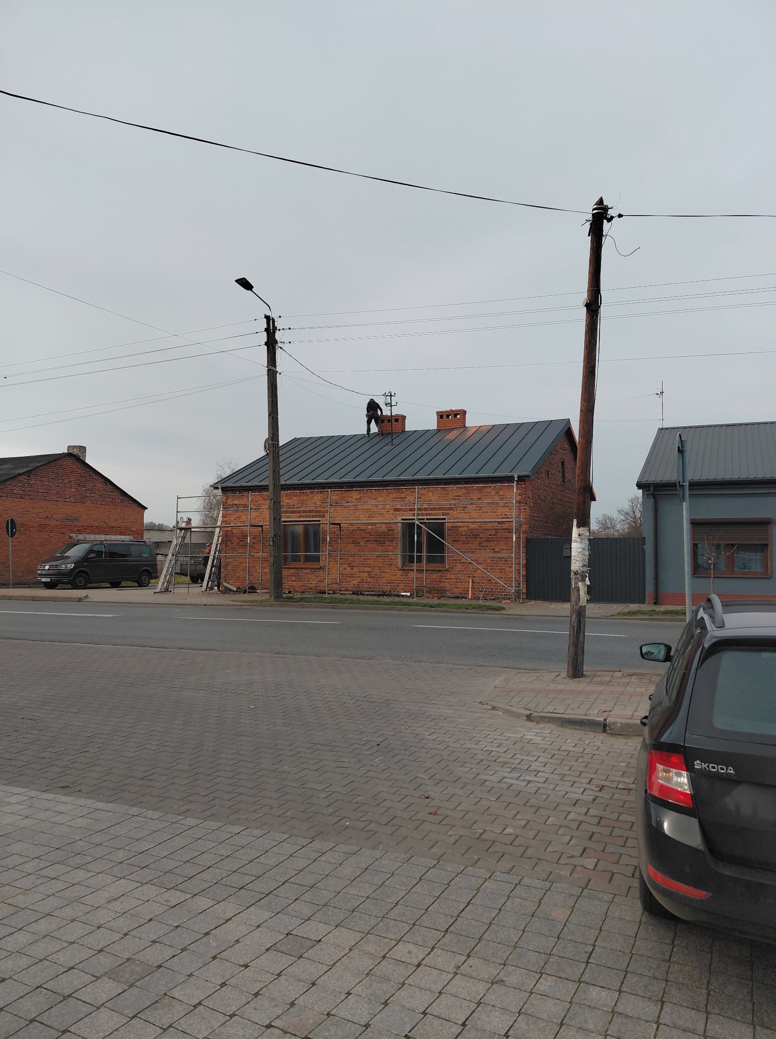 Ceglany budynek z nowym, ciemnym dachem w trakcie prac. Na dachu widoczna sylwetka pracownika. Rusztowania przy ścianie. Ujęcie z poziomu ulicy, pochmurne niebo.