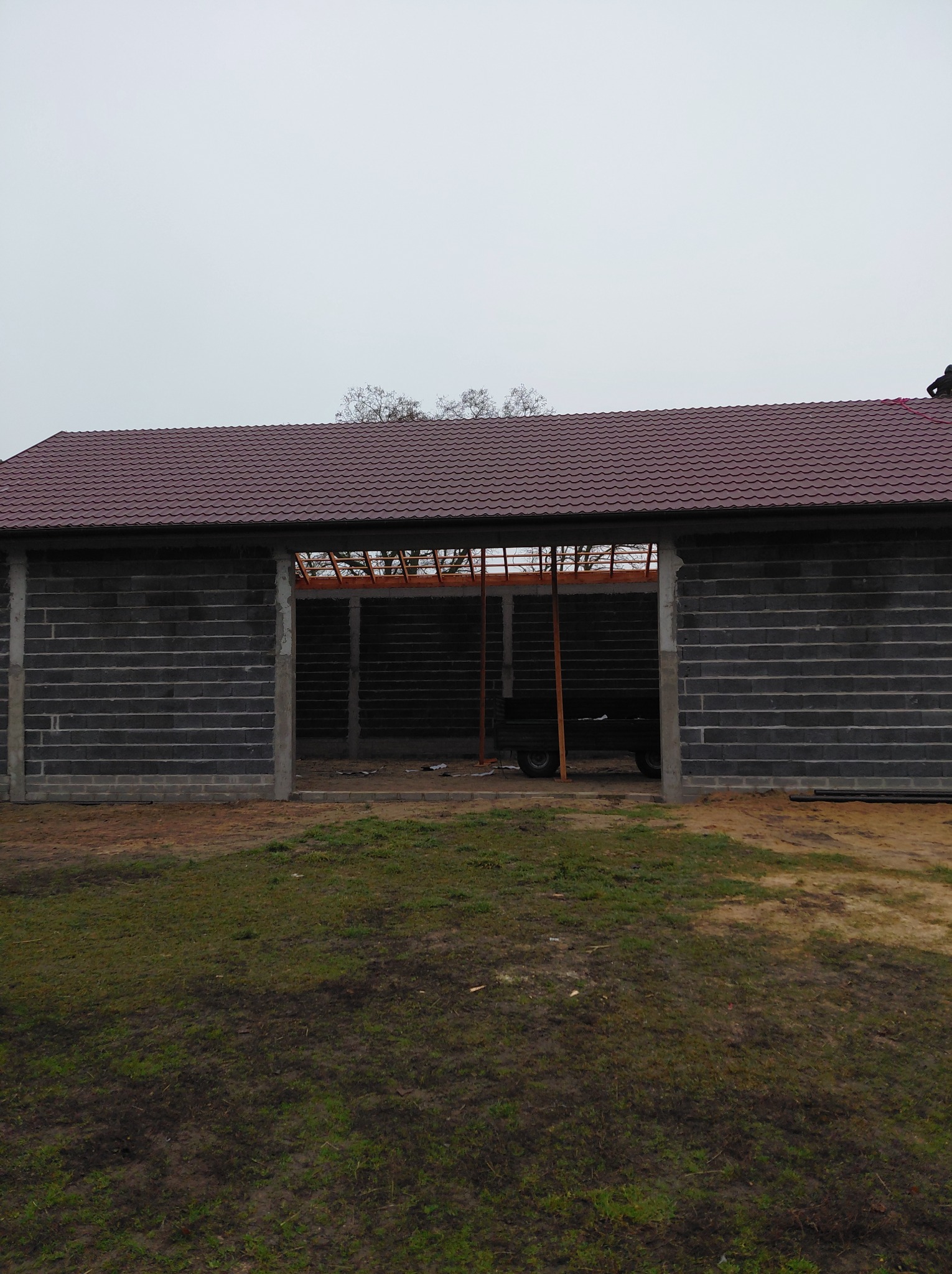 Budynek w trakcie budowy z ciemną dachówką, widoczna więźba dachowa i ściany z szarego bloczka. Na dachu widoczna osoba w trakcie prac.