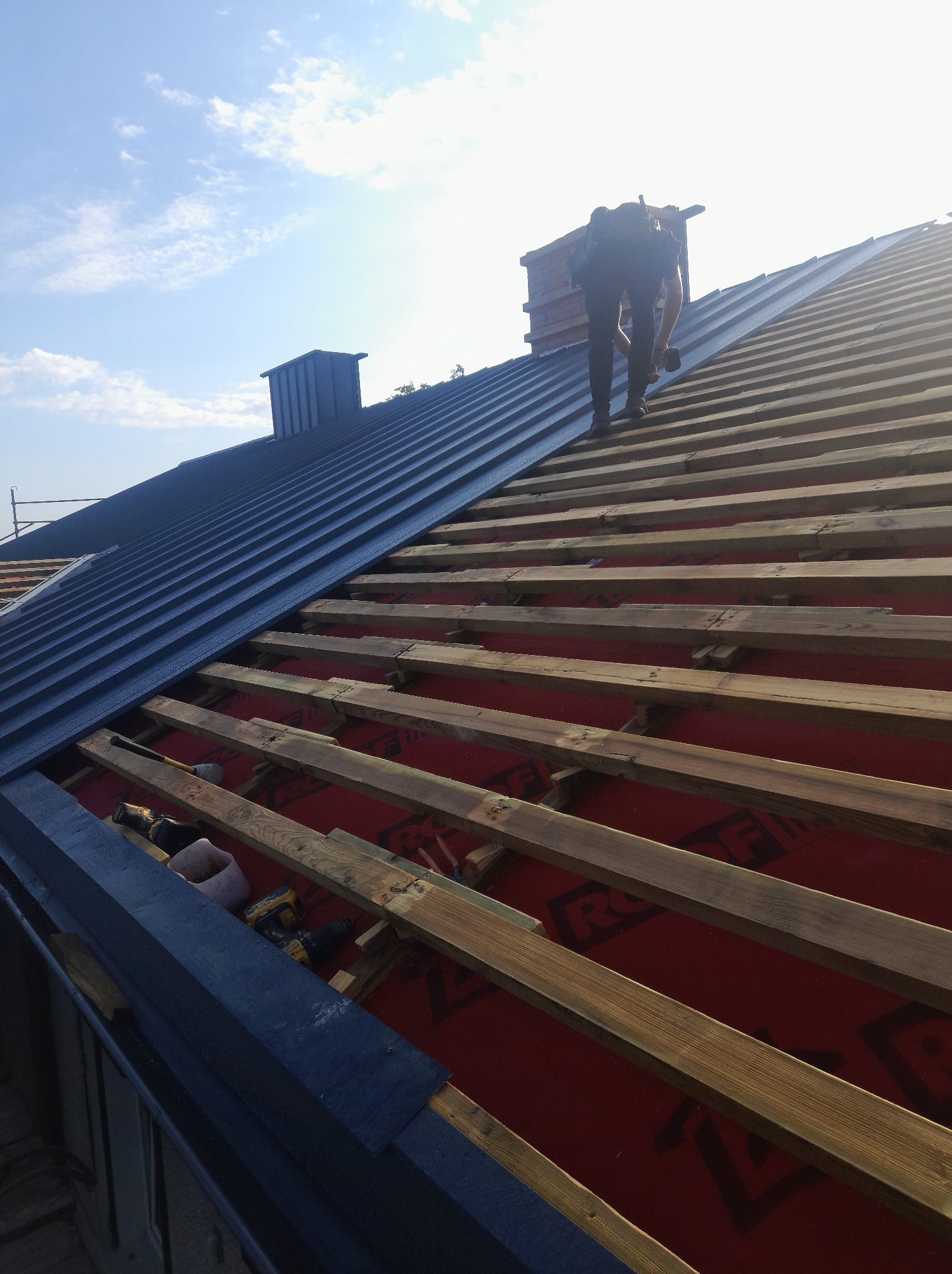 Montaż ciemnoszarego dachu z blachy na drewnianej konstrukcji. Widoczny komin i narzędzia pracy. Praca na wysokości w słoneczny dzień.