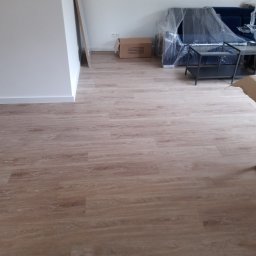 Usługi wykończeniowe - Jasne panele podłogowe w nowym mieszkaniu, meble zabezpieczone folią, kartonowe pudło i białe ściany.