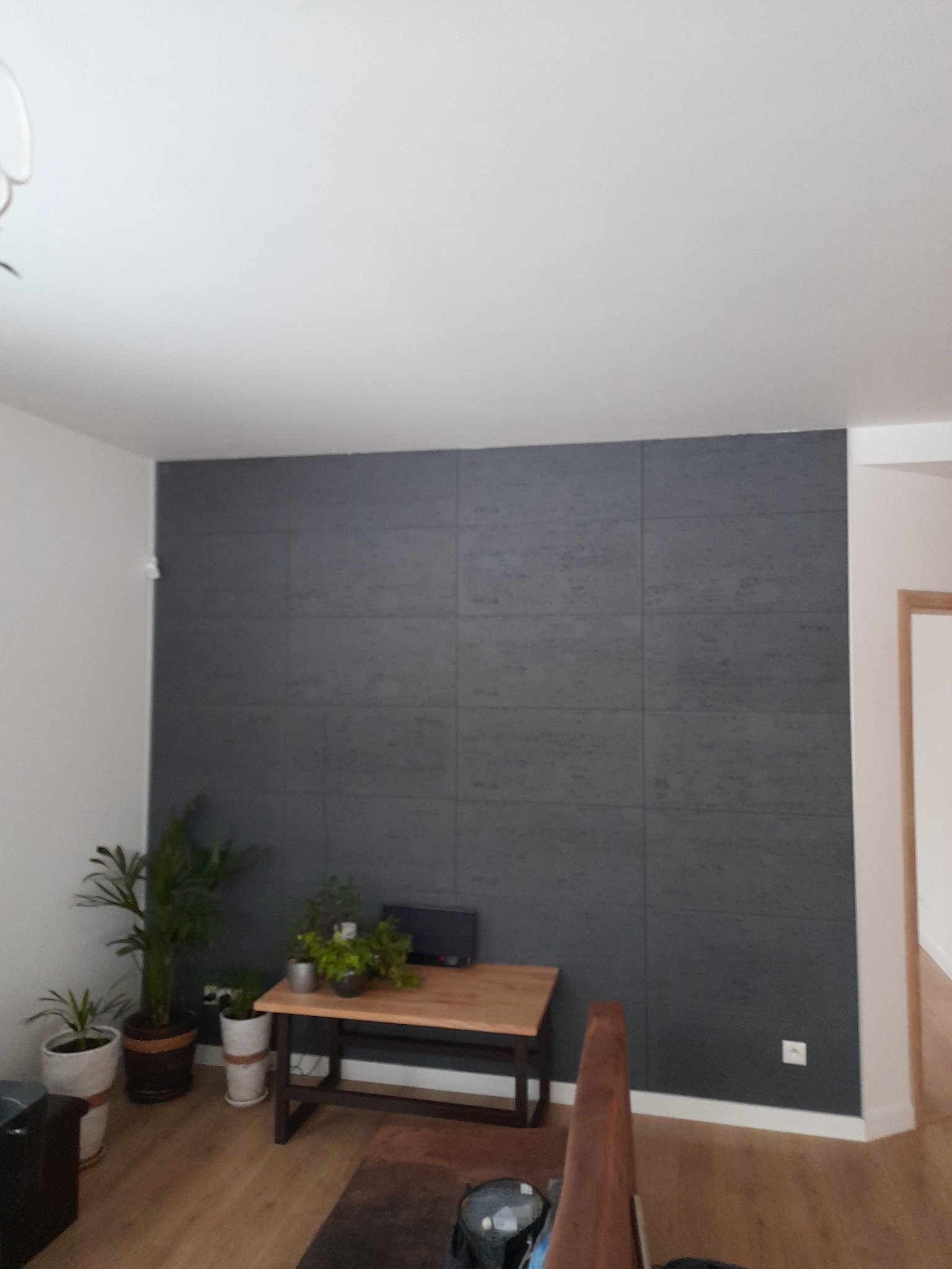 Szara ściana wykończona panelami dekoracyjnymi w salonie, z drewnianym stolikiem i roślinami doniczkowymi, widok z perspektywy schodów.