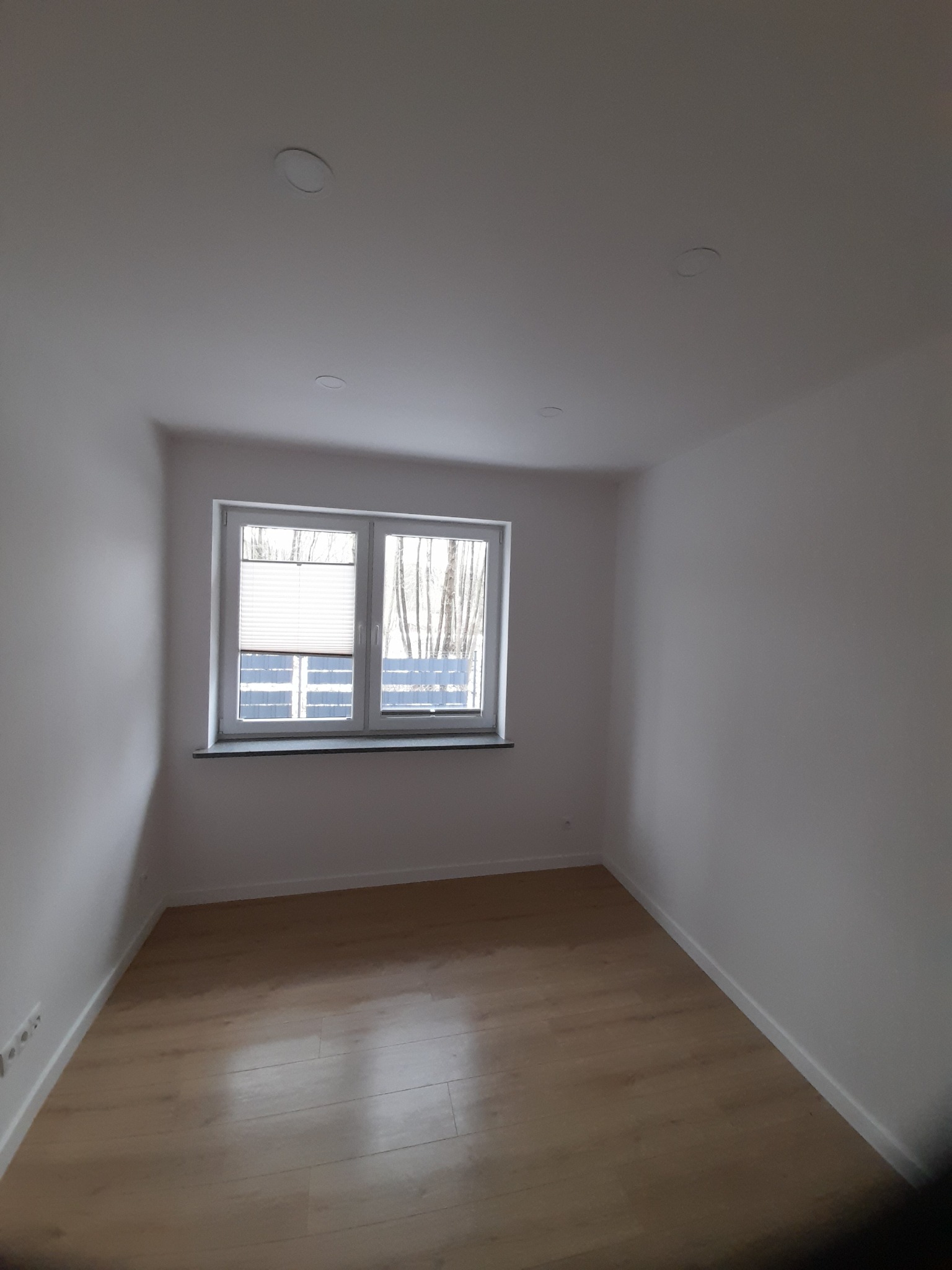 Pusty, odnowiony pokój z parkietem, białymi ścianami, oknem z żaluzjami plisowanymi i dwoma wbudowanymi punktami świetlnymi w suficie.