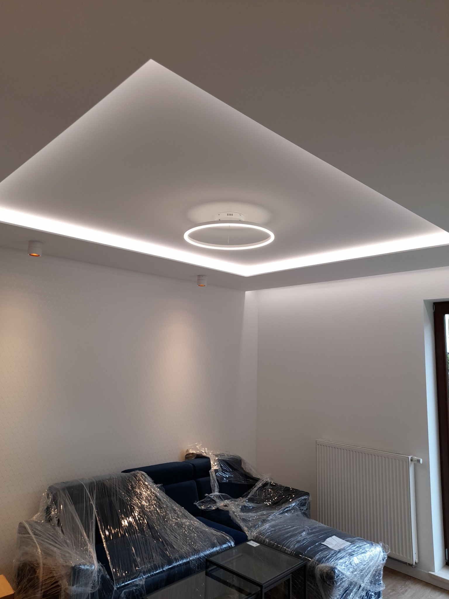 Nowoczesny sufit podwieszany z oświetleniem LED w kształcie kwadratu i okręgu, zabezpieczone meble folią ochronną.