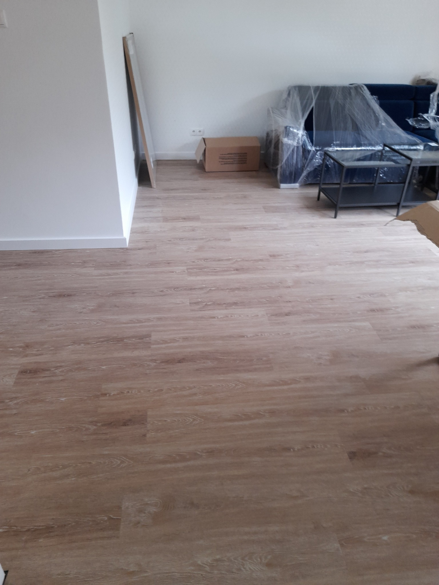 Jasne panele podłogowe w nowym mieszkaniu, meble zabezpieczone folią, kartonowe pudło i białe ściany.