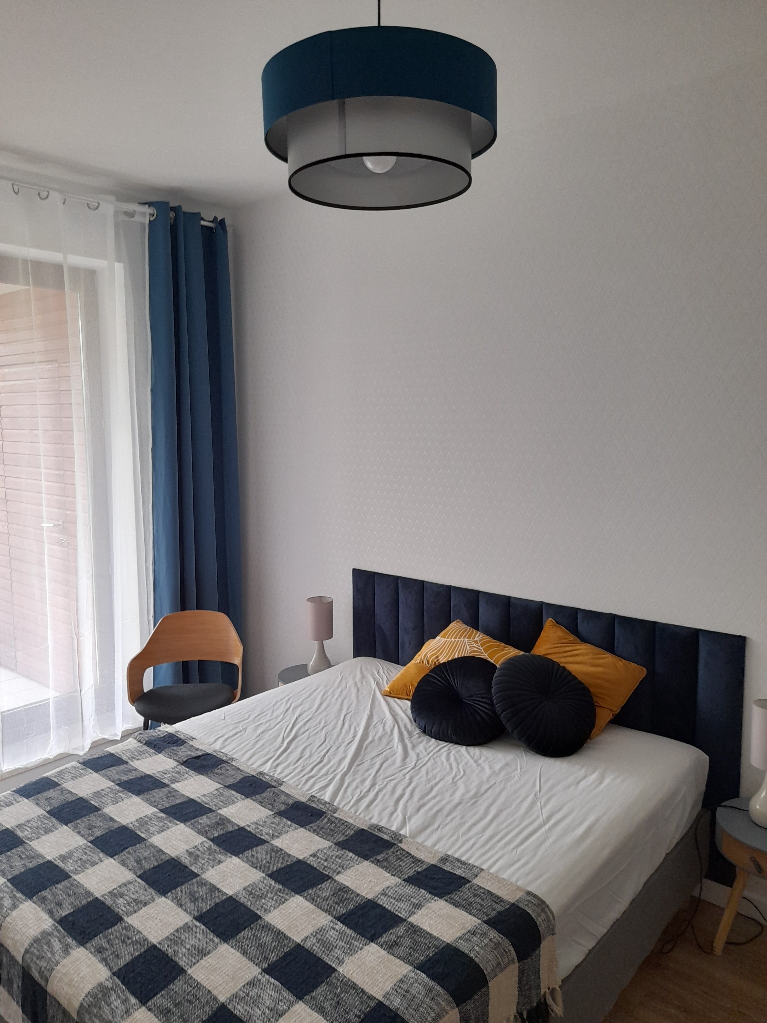 Wykończona sypialnia w Kielcach z łóżkiem z pikowanym, granatowym zagłówkiem, nakrytym białą pościelą, żółtymi poduszkami i pledem w kratę, drewnianym krzesłem oraz lampą sufitową z granatowym...