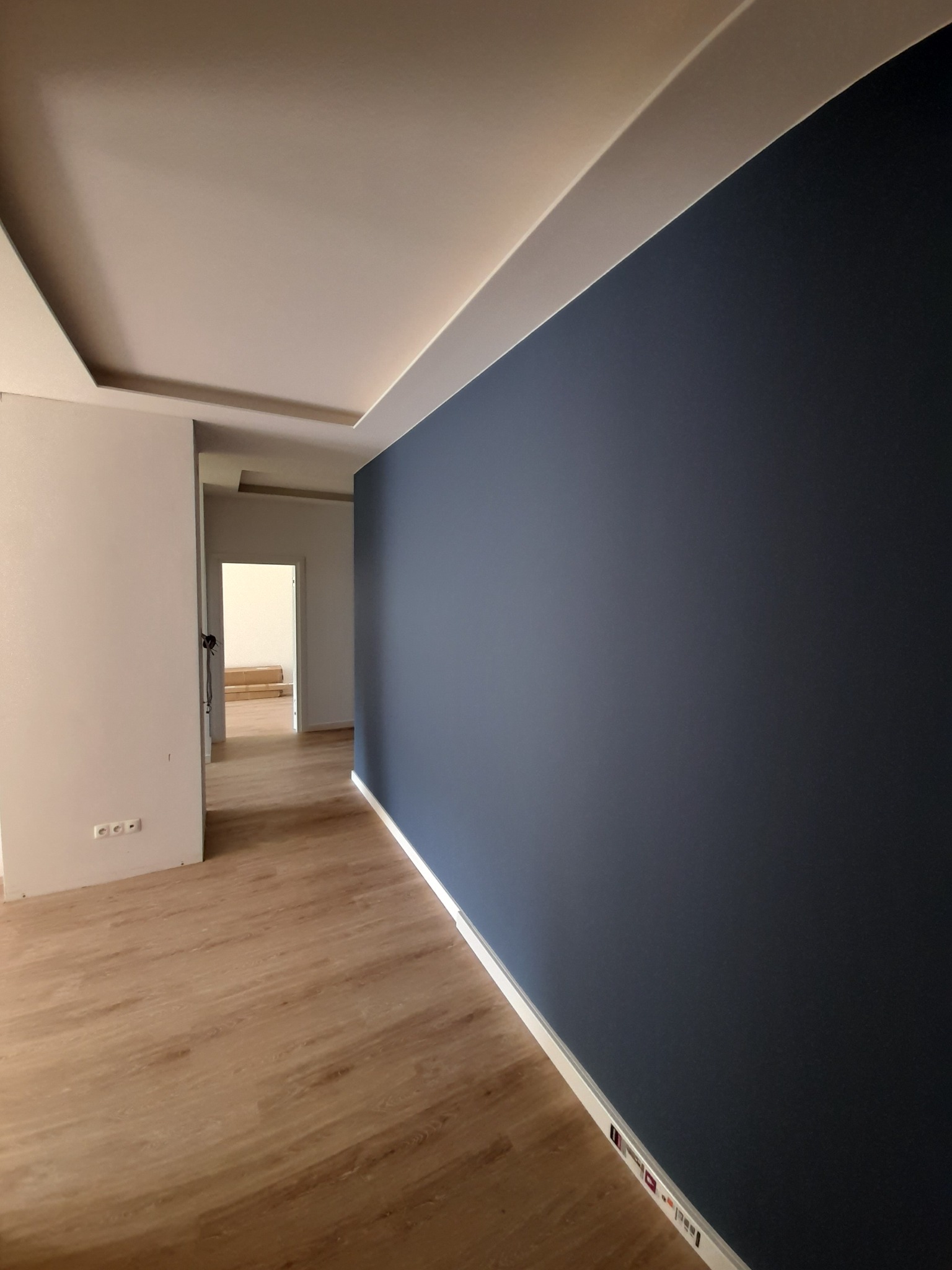 Wnętrze po remoncie: ściana pomalowana na granatowo, drewnopodobna podłoga, biały sufit z oświetleniem LED, widok na kolejne pomieszczenie z kartonami na podłodze.