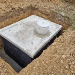 Betonowy zbiornik, prawdopodobnie szambo, umieszczony w wykopie z widocznymi ścianami ziemi i pokrywą z włazem. Widoczne stalowe uchwyty na pokrywie.
