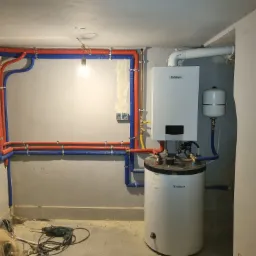 Biały piec gazowy Vaillant z podłączonymi rurami w izolacji czerwono-niebieskiej, widoczne narzędzia i pies leżący na podłodze w trakcie instalacji.