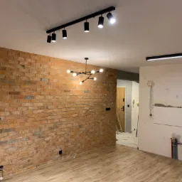 Wnętrze w trakcie remontu: ściana z cegły, drewniana podłoga, sufit z oświetleniem szynowym i nowoczesną lampą wiszącą, fragment ściany przygotowany pod glazurę z widocznymi rurami.