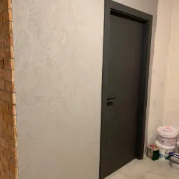 Ściana z efektem dekoracyjnym obok ceglanej kolumny, czarne drzwi z minimalistyczną klamką, widoczne puszki z farbą przy podłodze.