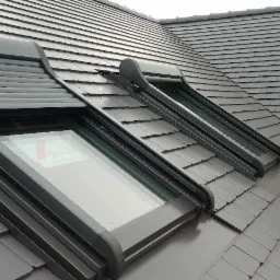 Szary dach z dachówki ceramicznej z dwoma oknami dachowymi Roto, jedno z roletą częściowo opuszczoną, drugie z roletą podniesioną.