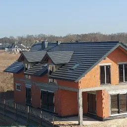 Dom w trakcie budowy z widocznym nowym, grafitowym pokryciem dachowym z dachówki ceramicznej, okna dachowe zamontowane, ściany z czerwonego pustaka.