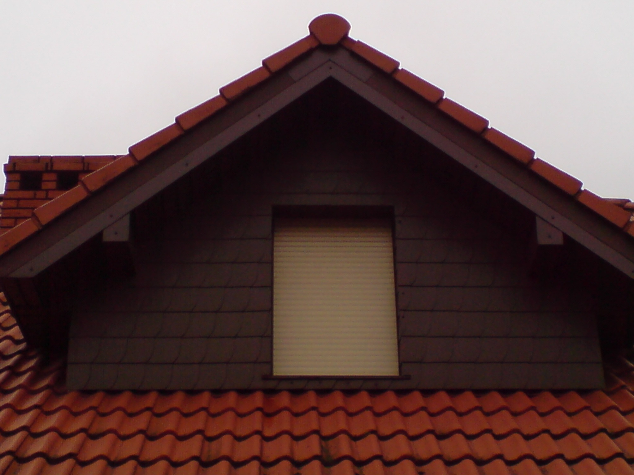 Szczyt dachu z oknem w lukarnie, pokryty czerwoną dachówką ceramiczną, z kominem w tle i szarą fasadą lukarny.