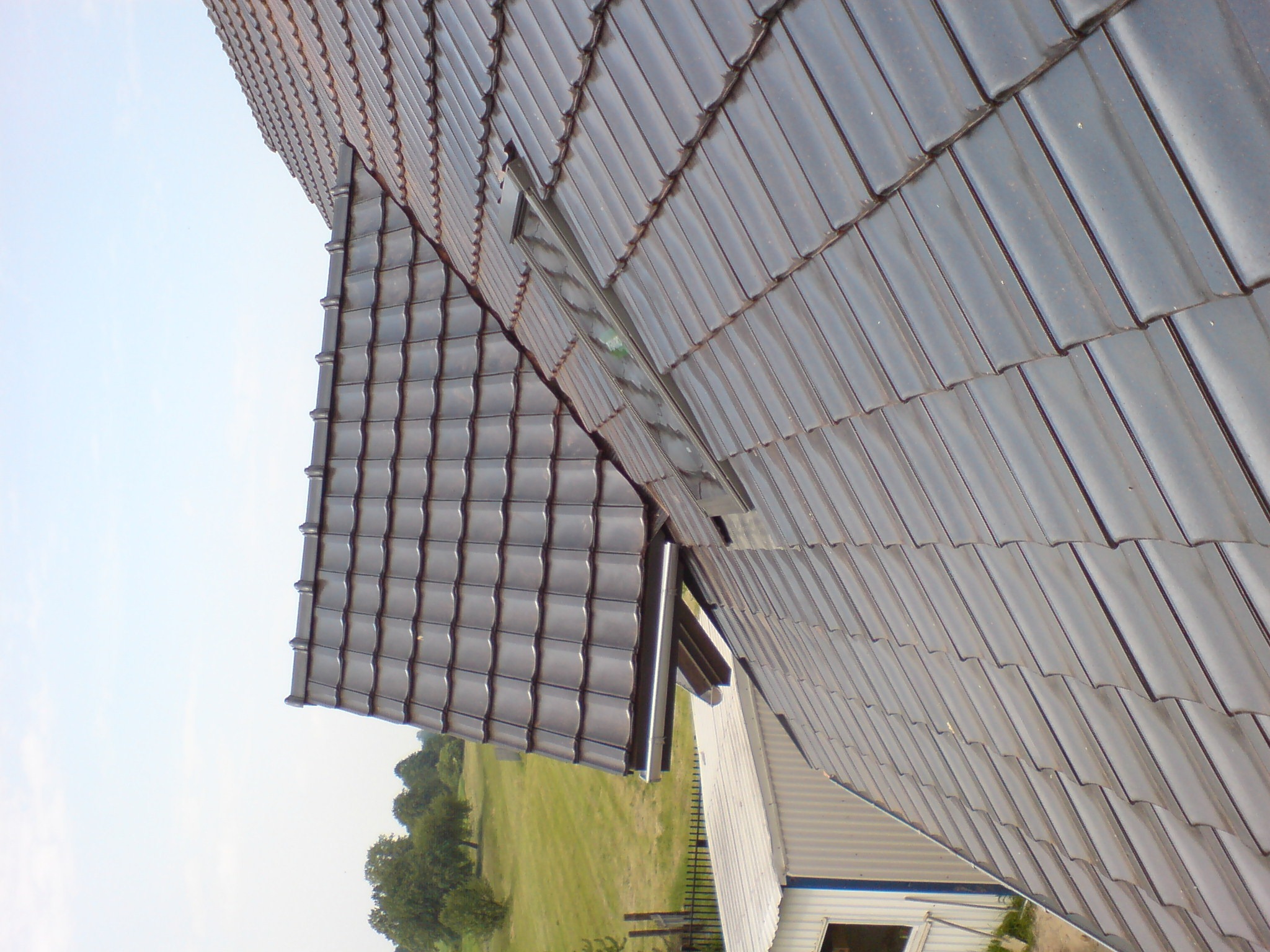 Widok z góry na fragment dachu z ciemnej dachówki ceramicznej, z widocznym oknem dachowym i rynną, na tle zielonego krajobrazu.