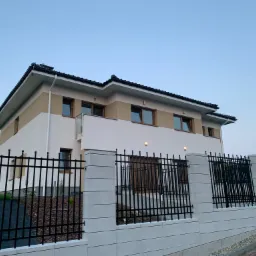 Nowoczesny dom jednorodzinny z elewacją w kolorach bieli i beżu, ogrodzony czarnym metalowym płotem na betonowej podmurówce z widocznym fragmentem podjazdu i samochodu w tle.