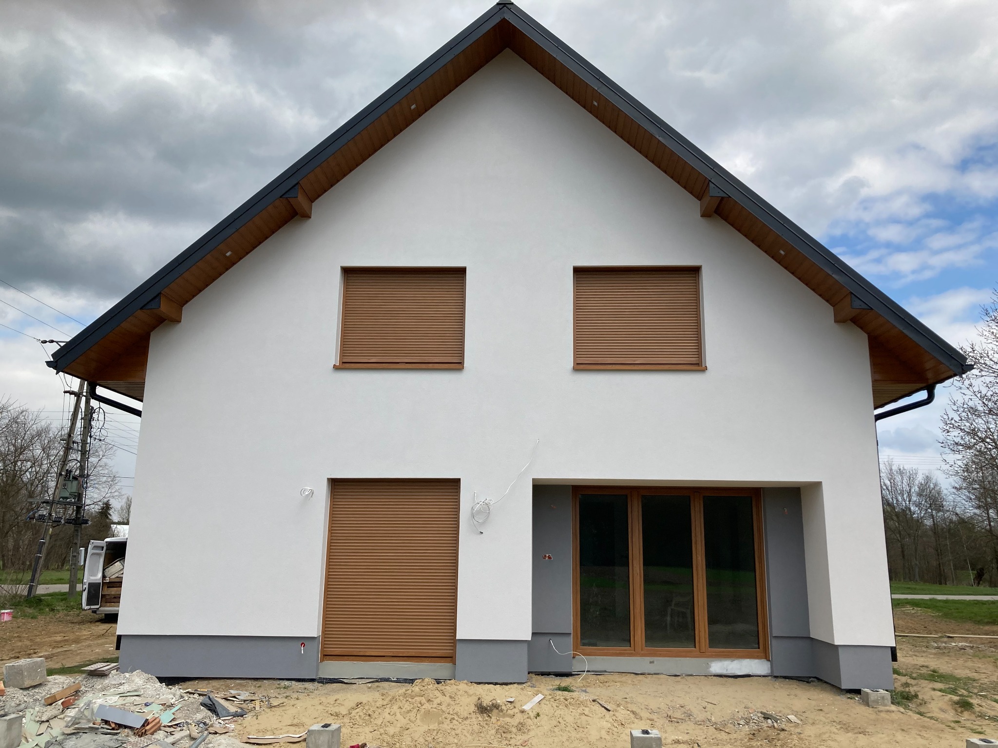 Nowoczesny dom jednorodzinny z białą elewacją, brązowymi roletami i szarą podmurówką w trakcie prac wykończeniowych, widoczne pozostałości materiałów budowlanych na gruncie.