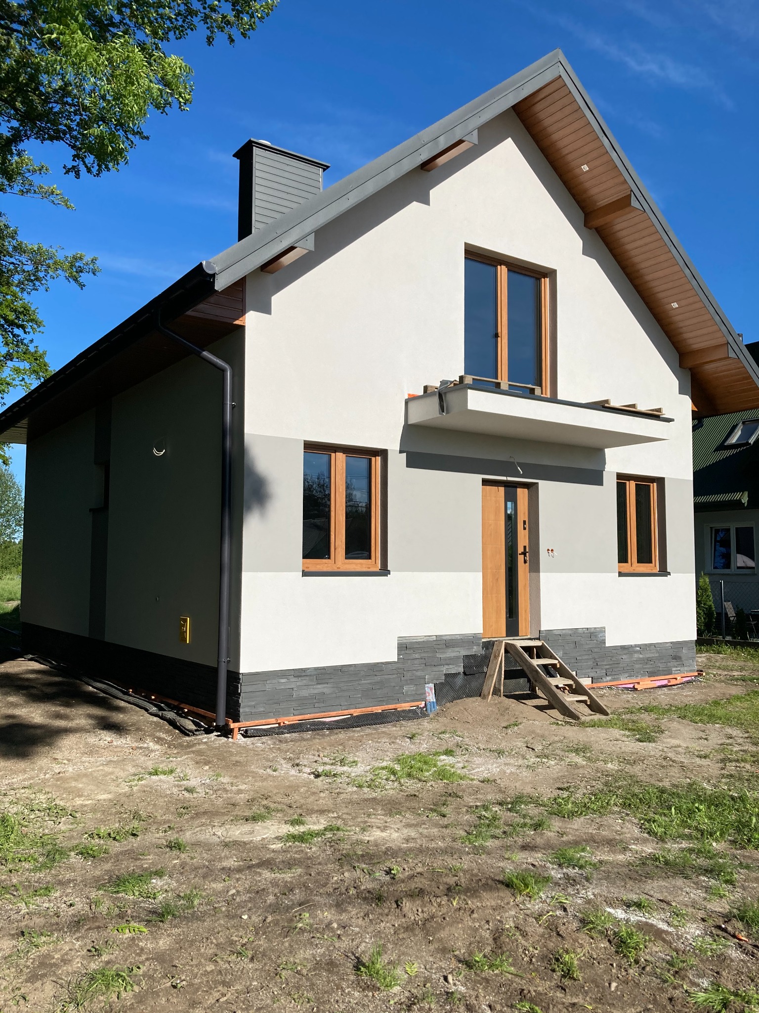 Nowoczesny dom jednorodzinny z elewacją w odcieniach szarości i bieli, drewniane okna i drzwi, widoczne przygotowania do wykończenia terenu wokół budynku, schodki wejściowe tymczasowe.