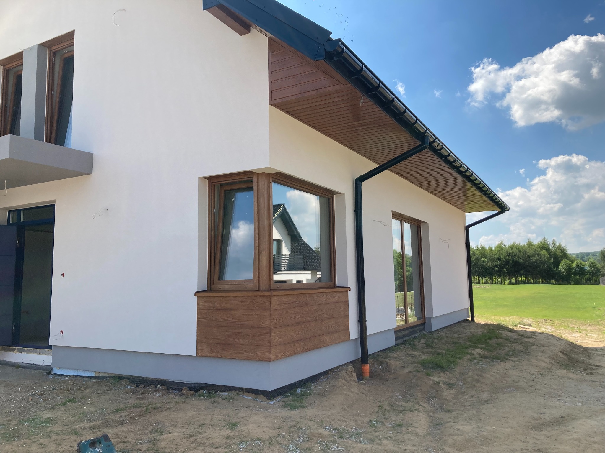 Nowoczesny dom z białą elewacją, brązowymi oknami i panelami imitującymi drewno, na tle błękitnego nieba z chmurami. Widoczne niedokończone prace ziemne wokół budynku.