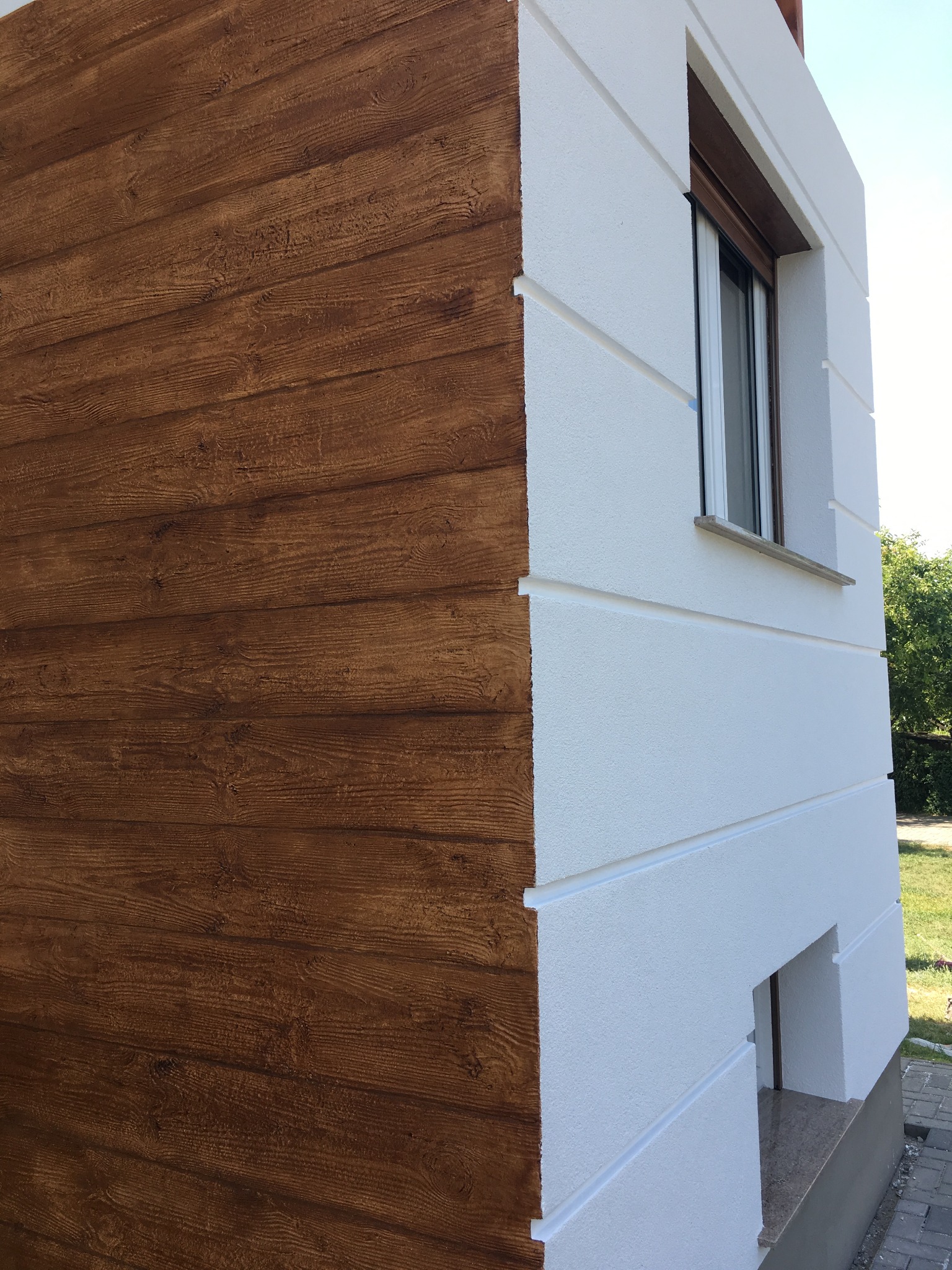 Fragment elewacji budynku z dwiema fakturami: biały tynk strukturalny i brązowe deski imitujące drewno.