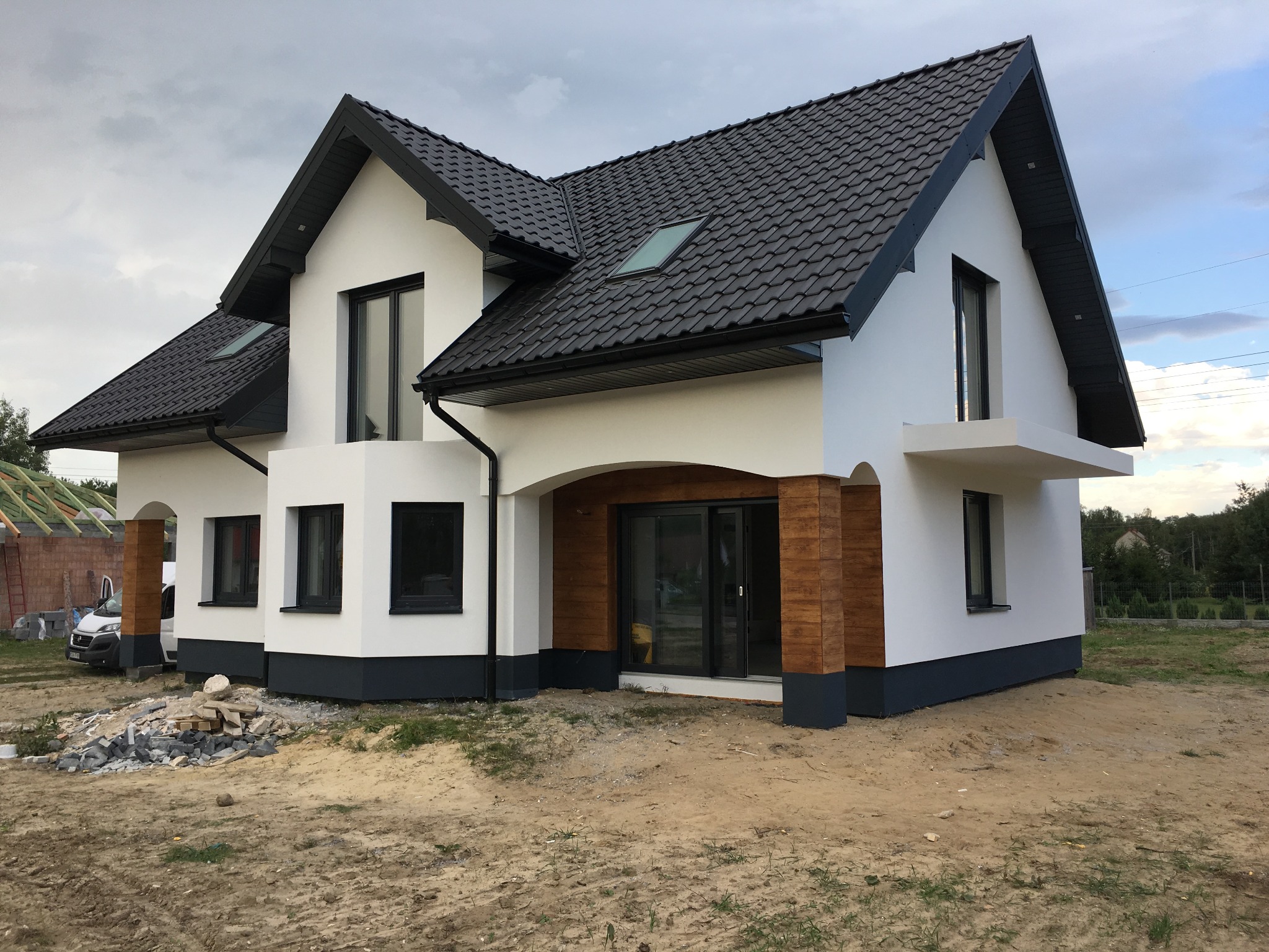 Nowoczesny dom jednorodzinny z białą elewacją, czarną dachówką i elementami dekoracyjnymi imitującymi drewno na kolumnach wejściowych, widoczny na tle pochmurnego nieba i niezagospodarowanego terenu.