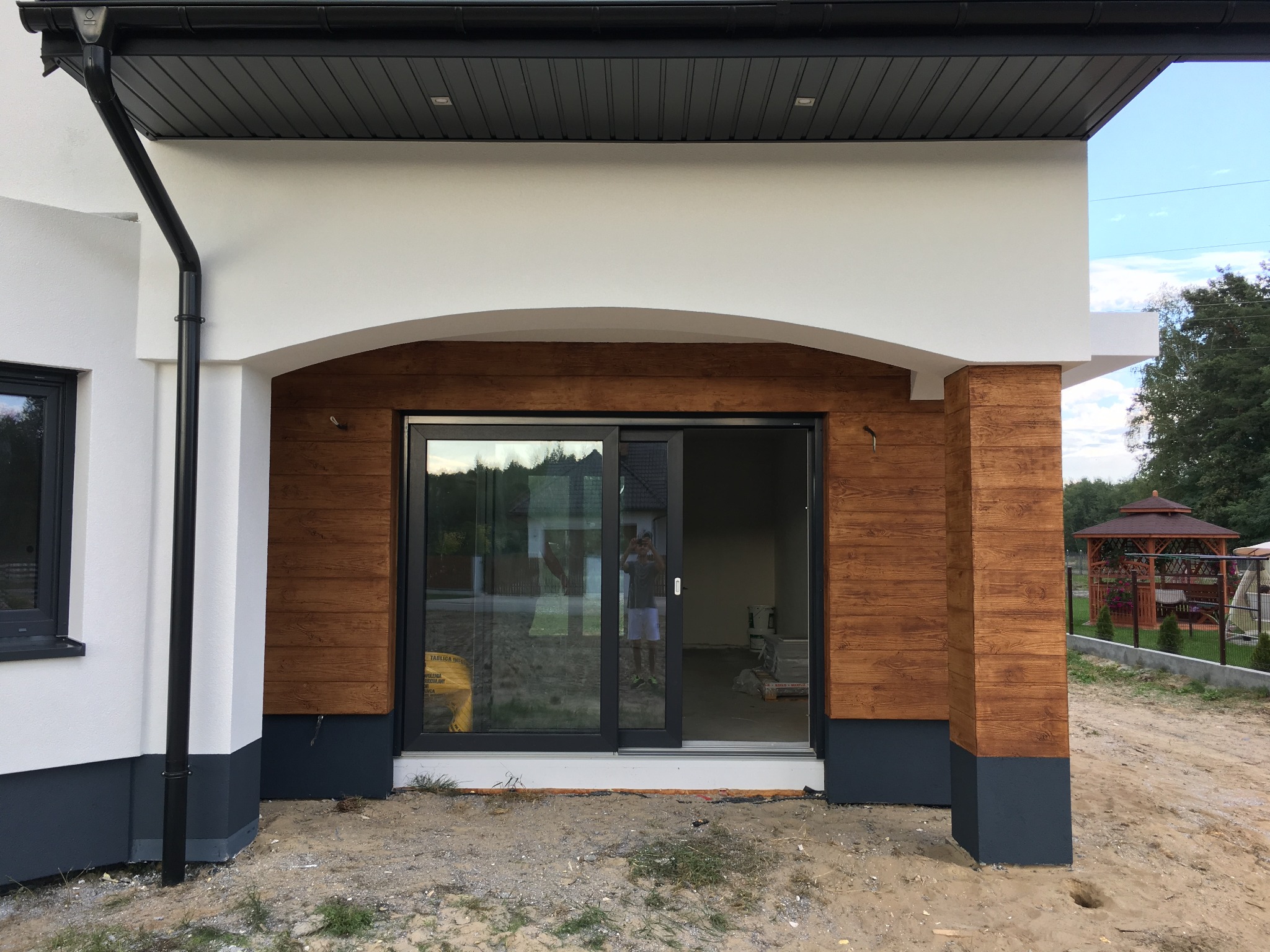 Nowoczesny dom z elewacją imitującą drewno, z czarną stolarką okienną i przesuwnymi drzwiami tarasowymi, widoczny fragment ogrodu z altaną.