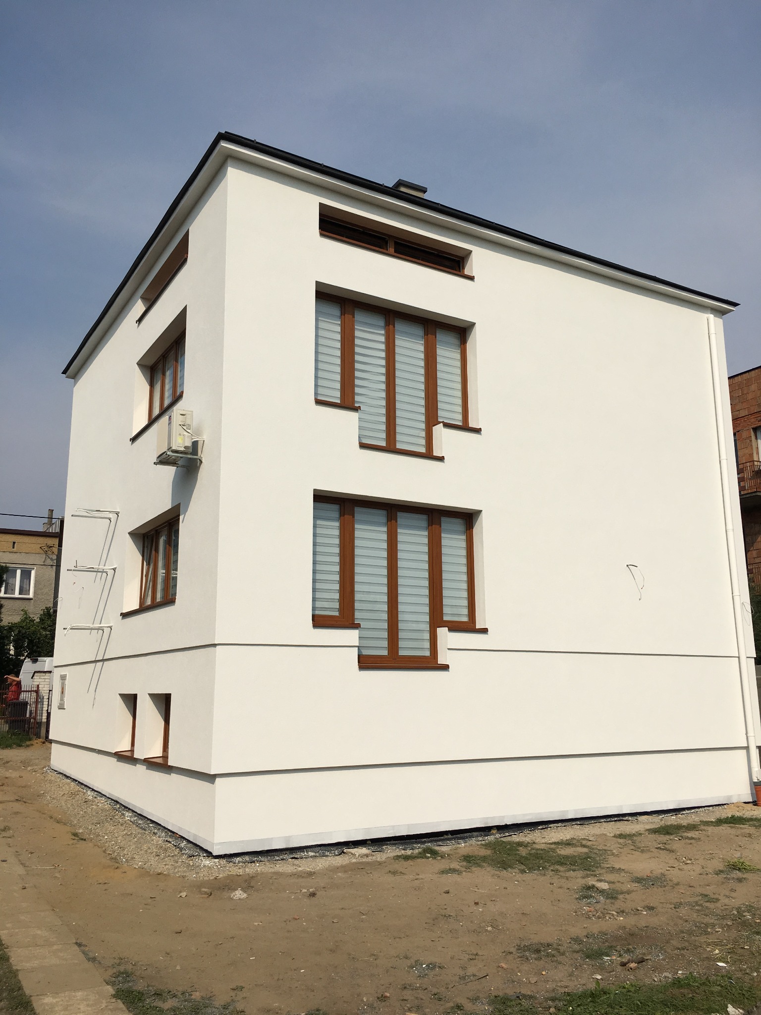 Nowoczesny, dwukondygnacyjny dom z białą elewacją, brązowymi ramami okiennymi i klimatyzatorem na ścianie bocznej, widoczny z dołu pod kątem.