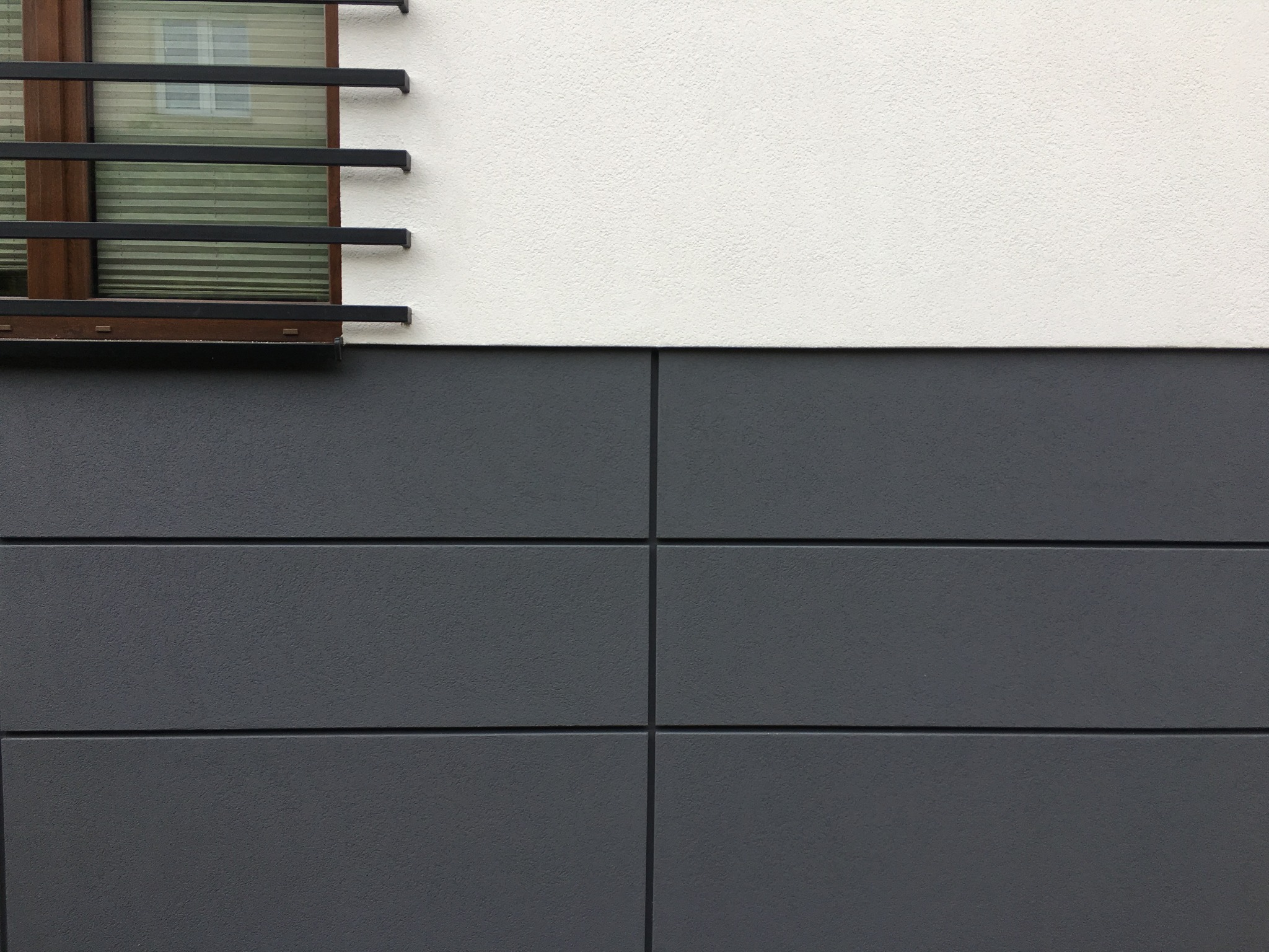 Fragment elewacji budynku z oknem zabezpieczonym metalową kratą. Górna część ściany jest biała, a dolna wykończona szarymi panelami.