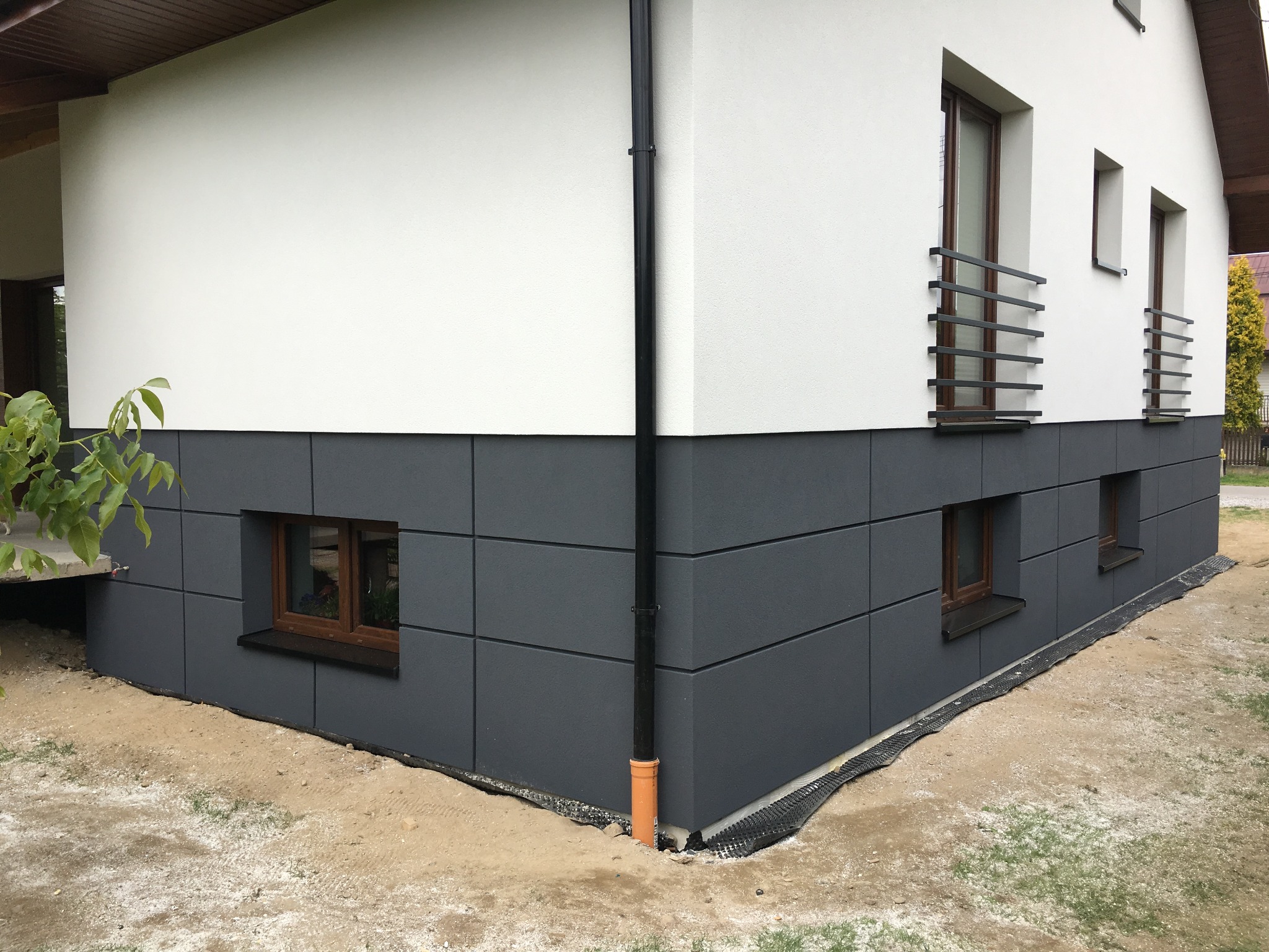 Wykończona elewacja domu z białym tynkiem górnym i szarymi panelami dolnymi, widoczne okna z brązowymi ramami i czarną rynną.