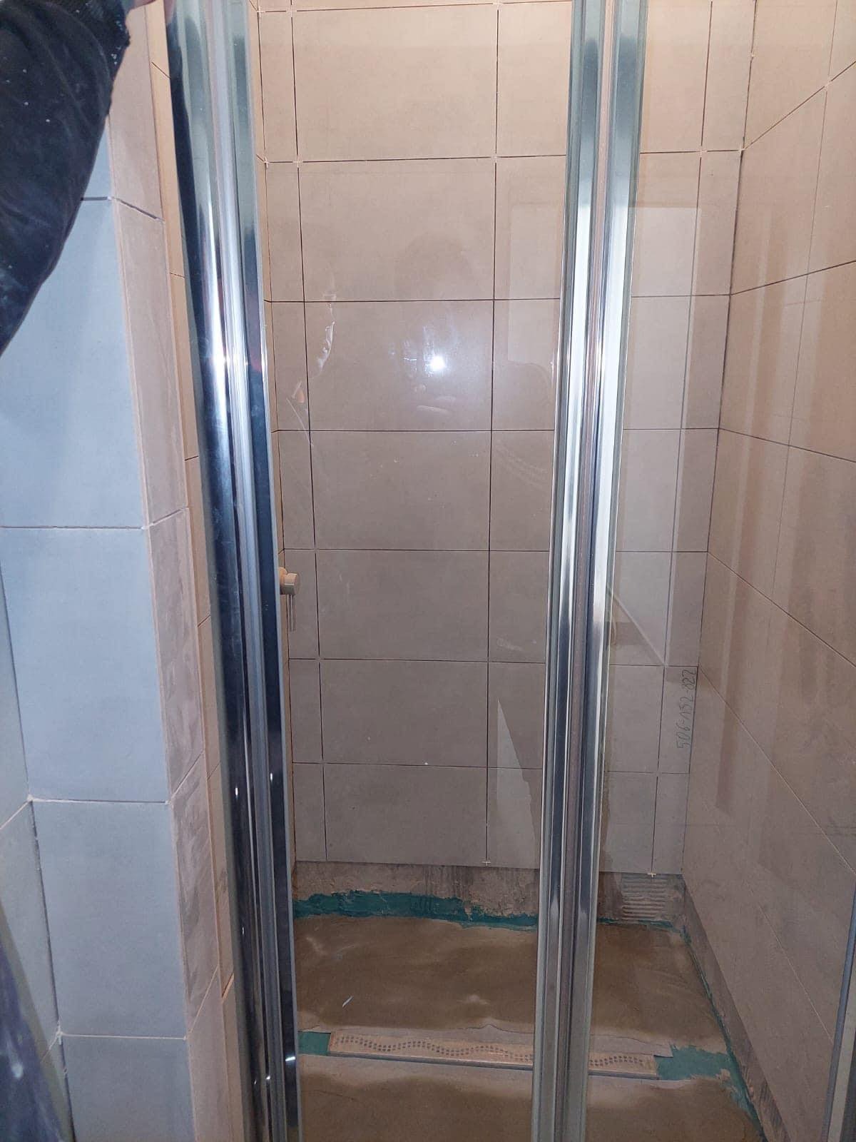 Montaż kabiny prysznicowej z chromowanymi profilami na tle beżowych płytek, widoczny surowy beton na podłodze i ślady prac wykończeniowych.