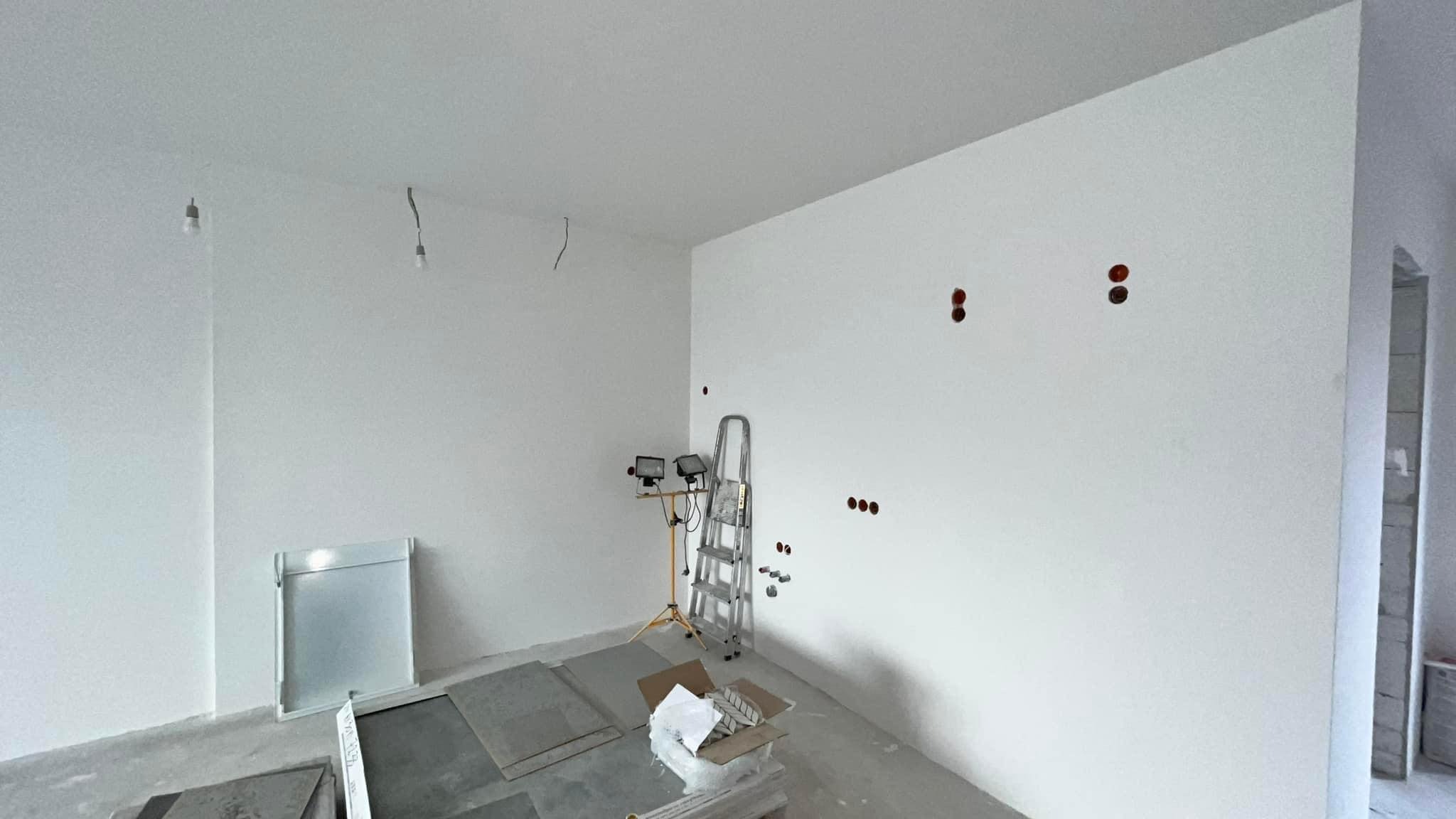 Wnętrze w trakcie malowania: białe ściany, widoczne puszki elektryczne, drabina aluminiowa, reflektor na statywie, płyty gipsowo-kartonowe i karton z elementami wykończeniowymi na podłodze, surowy...