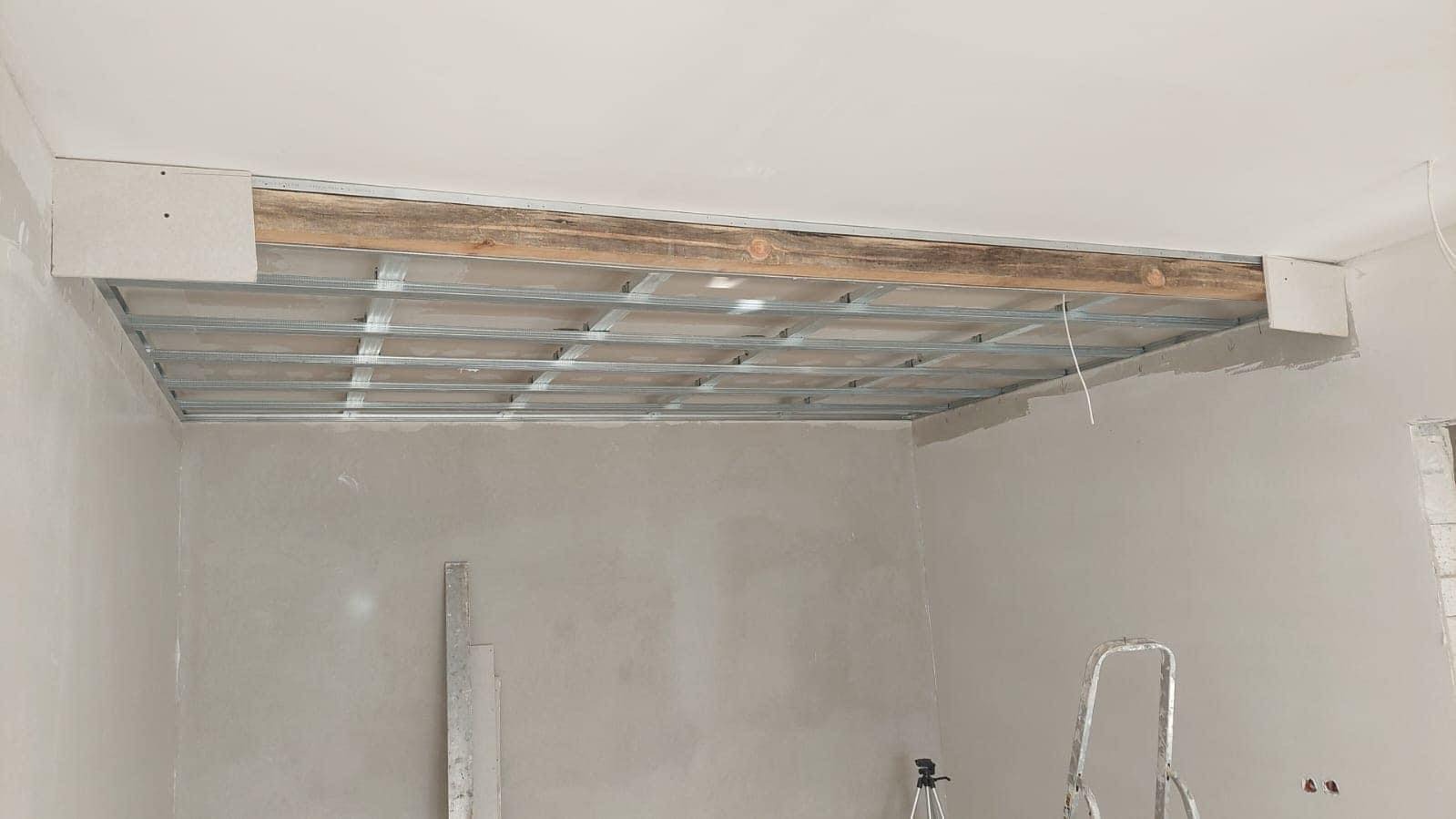 Metalowa konstrukcja sufitu podwieszanego, częściowo obudowana płytami gipsowo-kartonowymi, wsparta na drewnianej belce, widok z dołu na nieukończone wnętrze.
