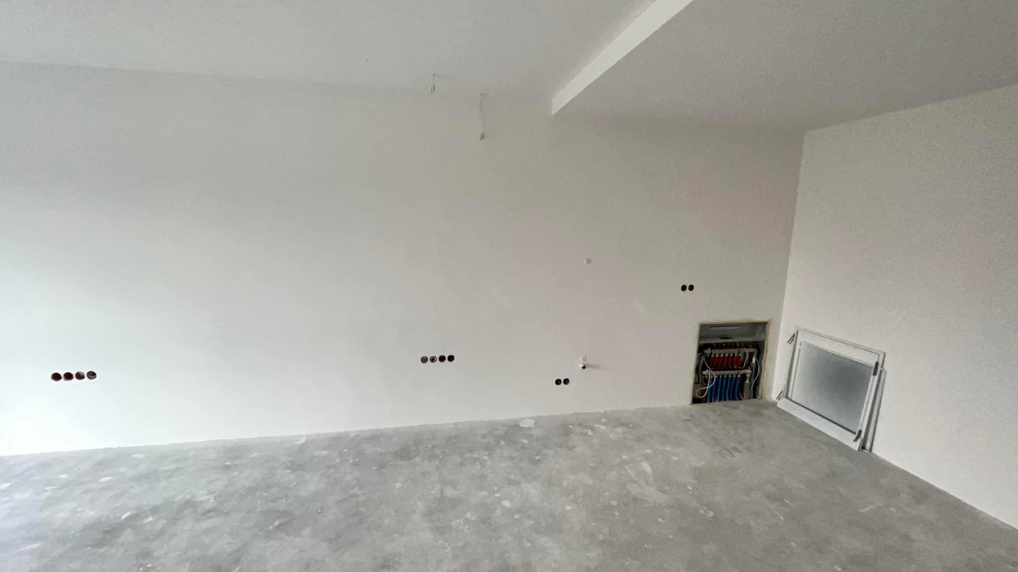 Surowe wnętrze w trakcie prac wykończeniowych, z widoczną instalacją centralnego ogrzewania w ścianie oraz przygotowanymi otworami na gniazdka elektryczne, świeżo wylana posadzka.