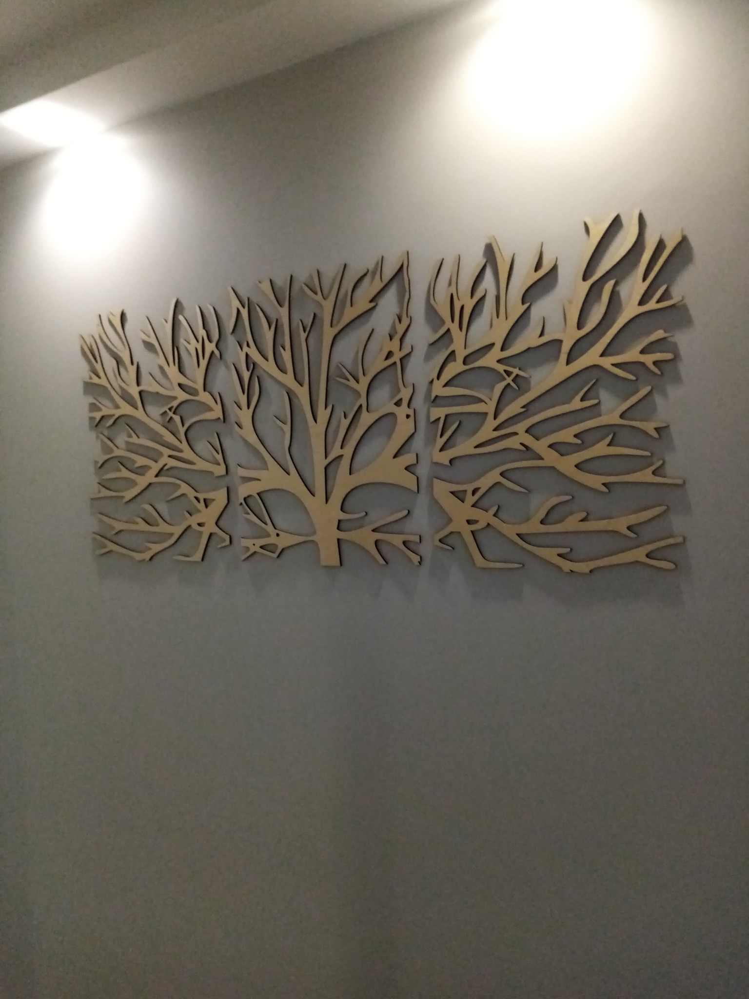 Dekoracja ścienna z motywem drzewa, wykonana z jasnego drewna, zamontowana na szarej ścianie pod dwoma punktowymi źródłami światła.